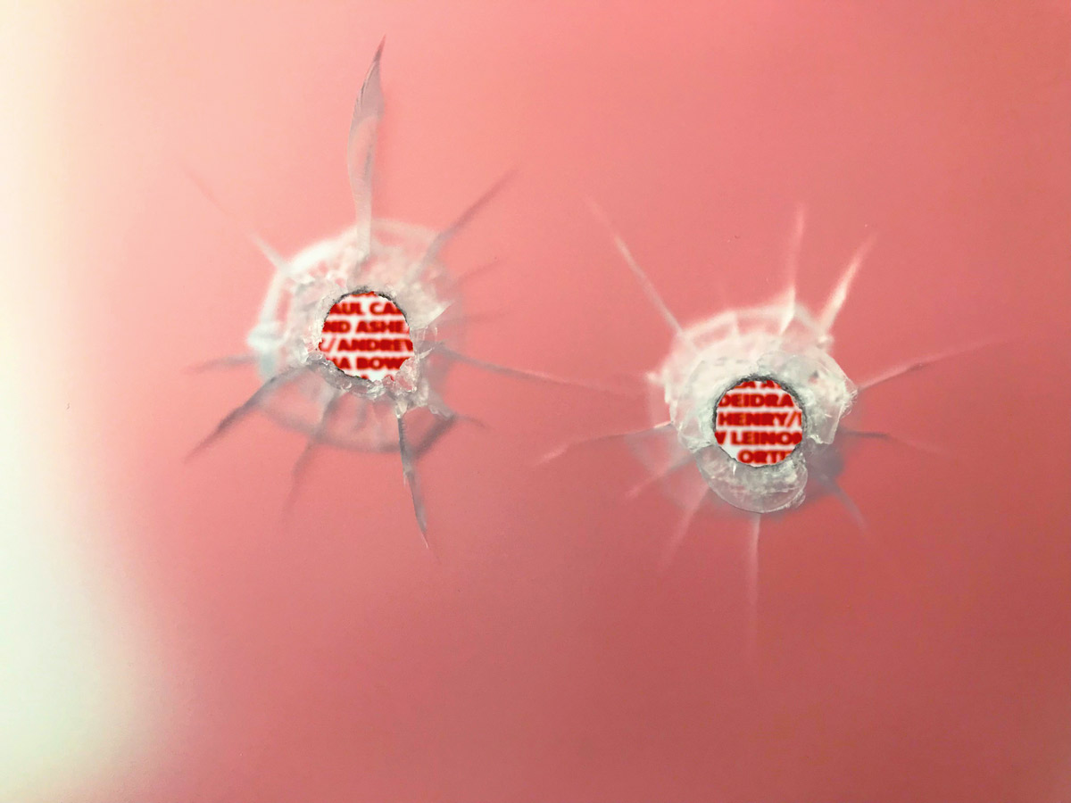 Close-up of two bullet holes. Through each, red names are sharp and readable.