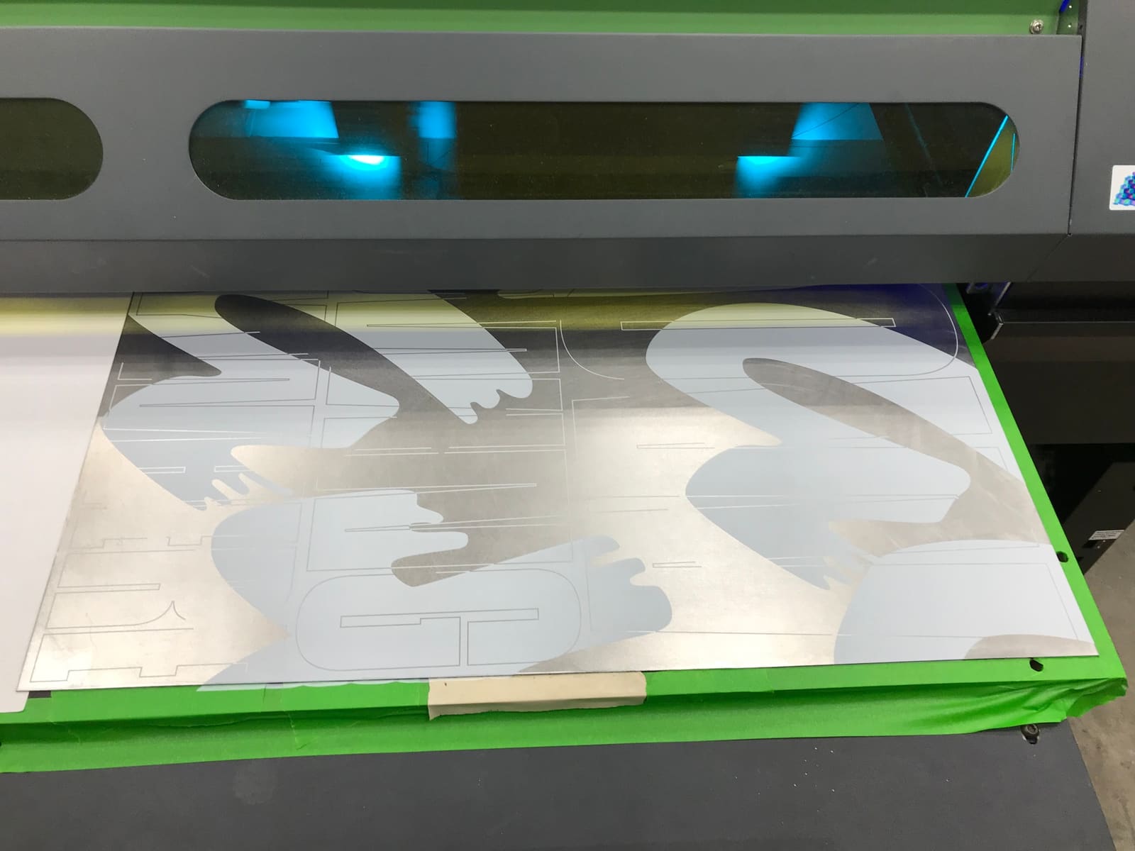 A steel plate on the bed of a large-format UV flatbed printer, with the white wordmark printed on it.
