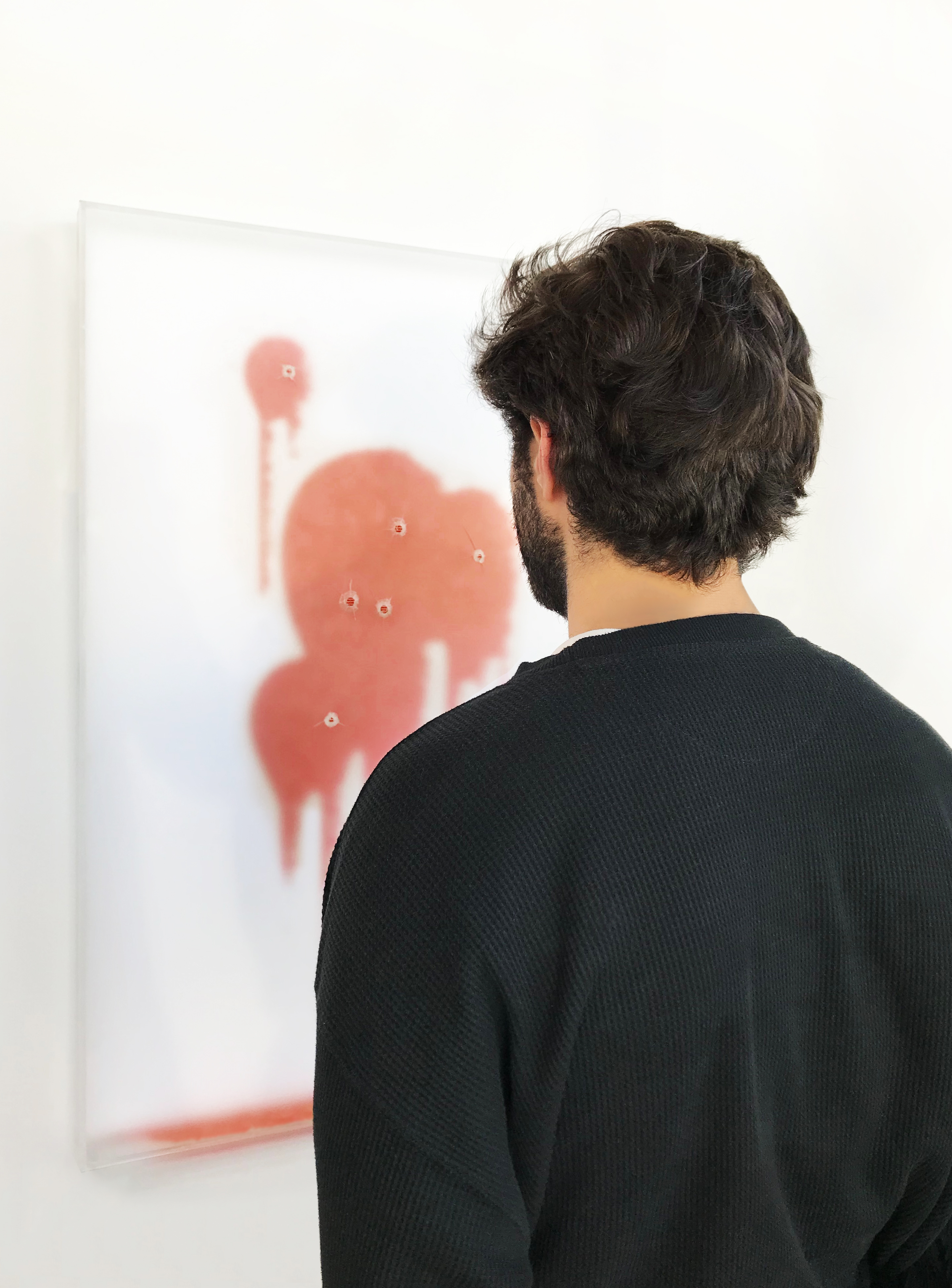 A man stands in front of the piece on a gallery wall, looking at the red stain behind the frosted acrylic and its bullet holes.