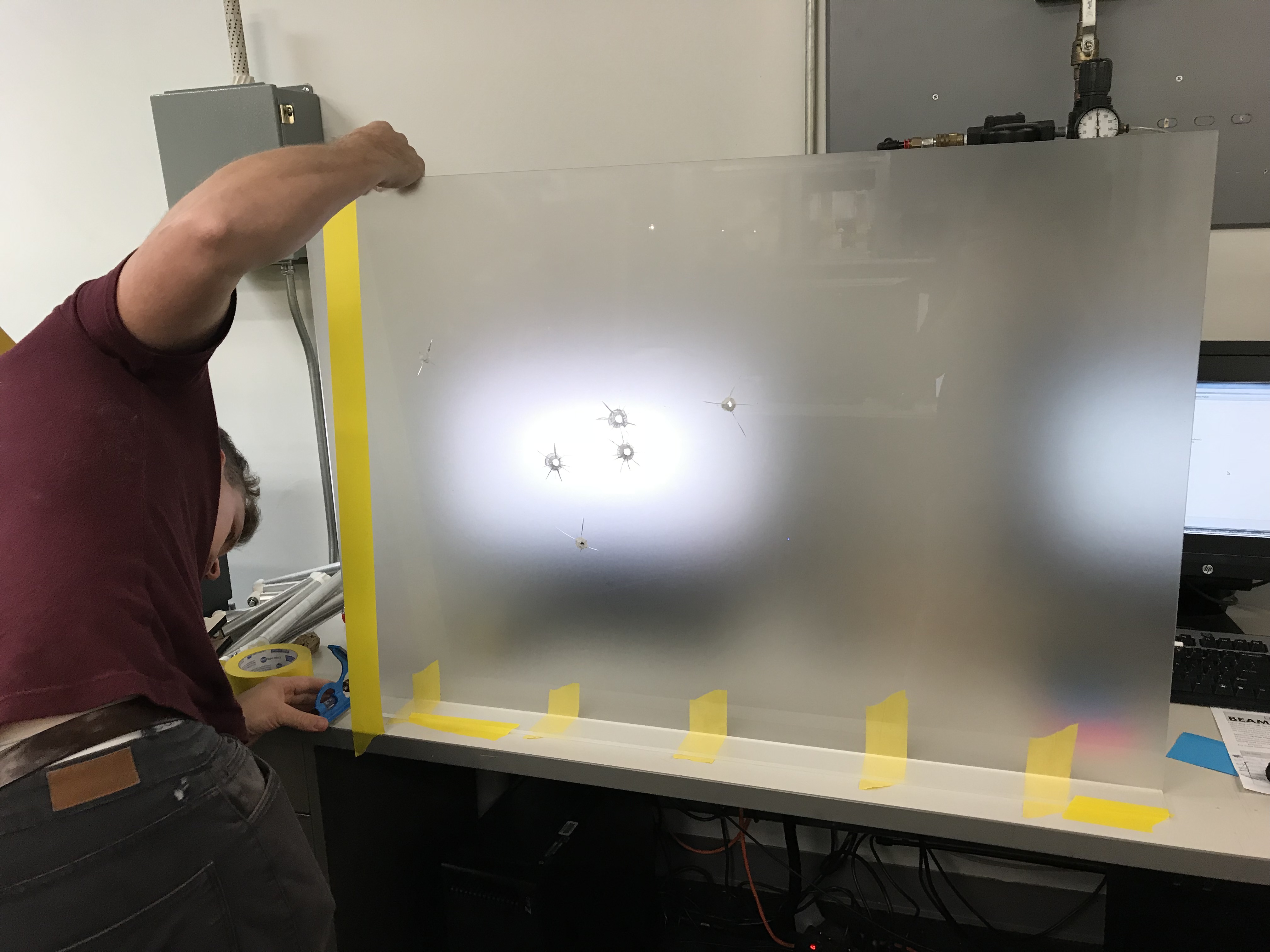 A person lifts a large frosted acrylic sheet into place; light shows through the bullet holes.