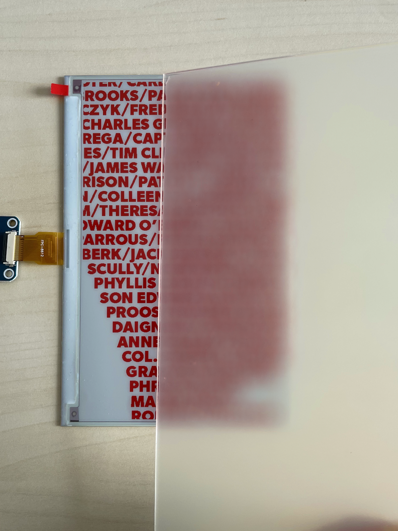 The e-paper display, half covered by frosted acrylic: sharp red names on one side, a soft red blur on the other.
