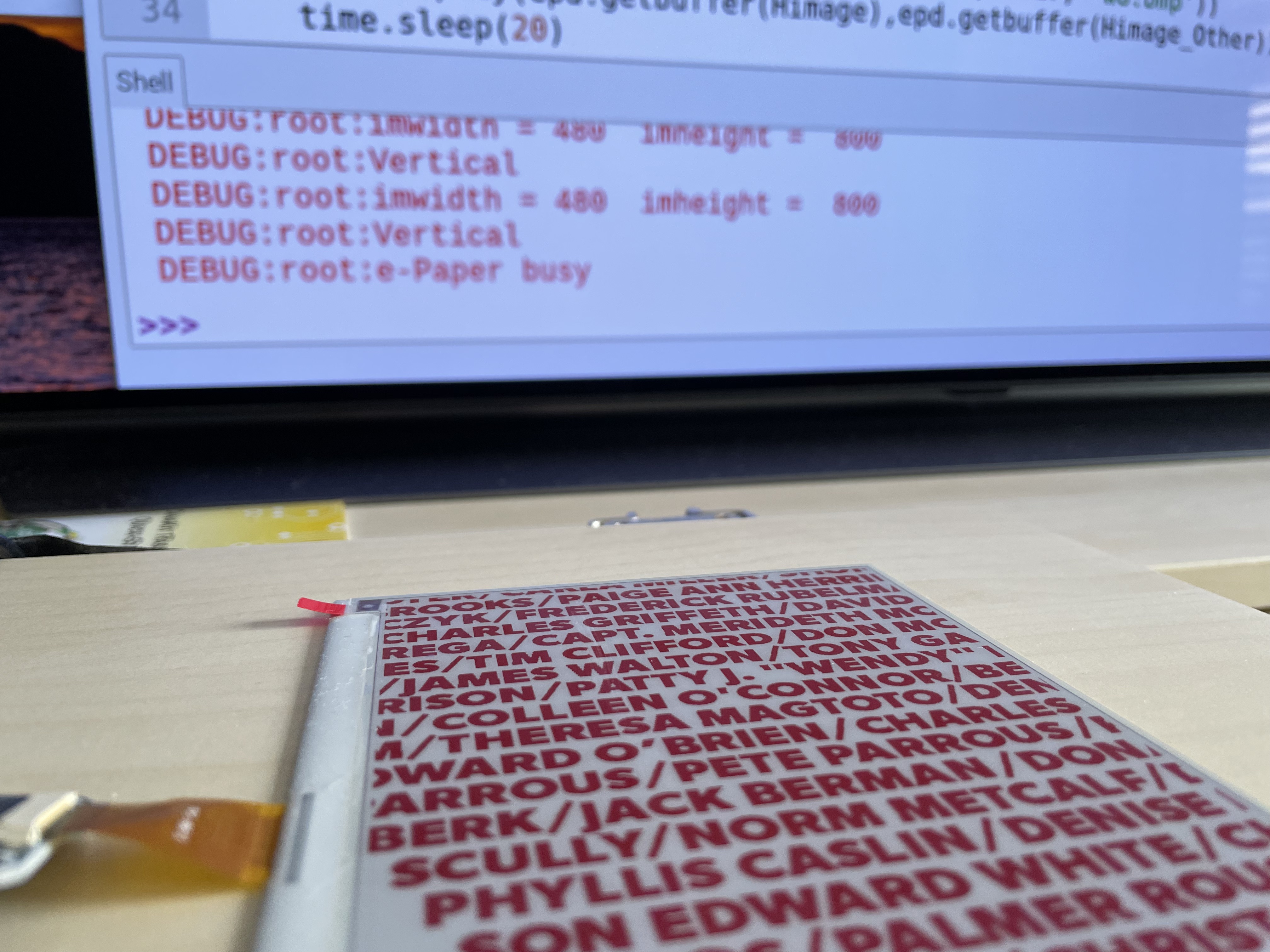 The e-paper display of names in front of a laptop showing the Python program’s debug output.
