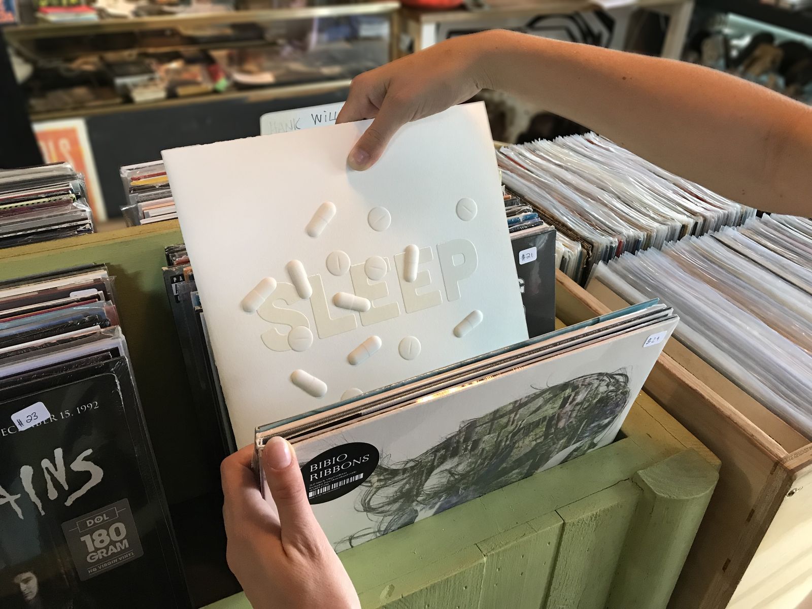 A hand pulls a white record sleeve titled Sleep, embossed with raised pill shapes, from a bin in a record store.