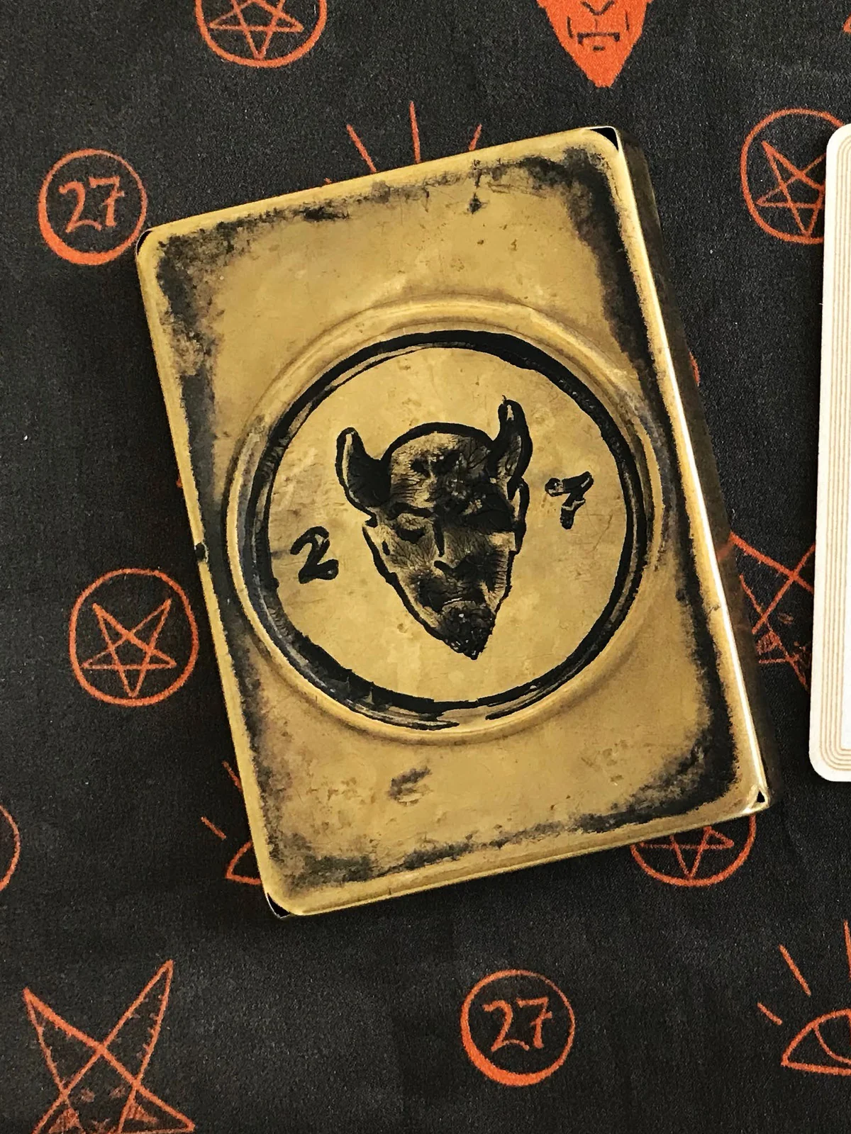 A worn brass case, darkened at the edges, stamped with a horned devil’s head between a 2 and a 7, lying on black silk printed with orange pentagrams and 27s.