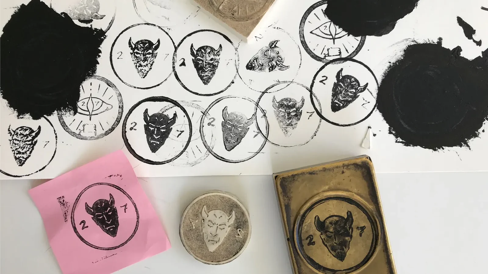 Test prints of the devil stamp in black ink, the laser-cut rubber stamp and the stamped brass case.