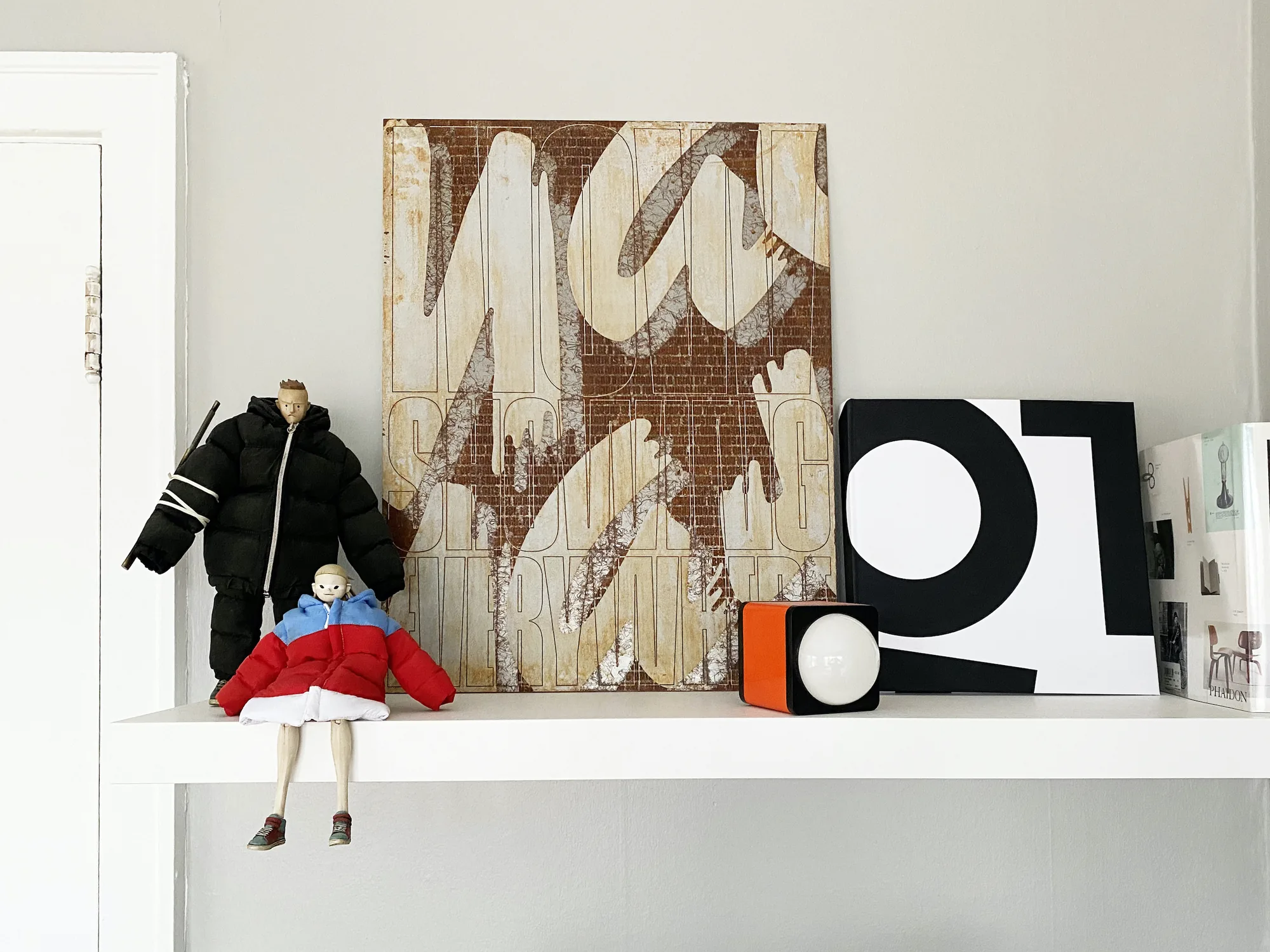 The rusted poster leaning against a grey wall on a white shelf, between two figurines in puffer jackets, an orange cube lamp and a black-and-white book.