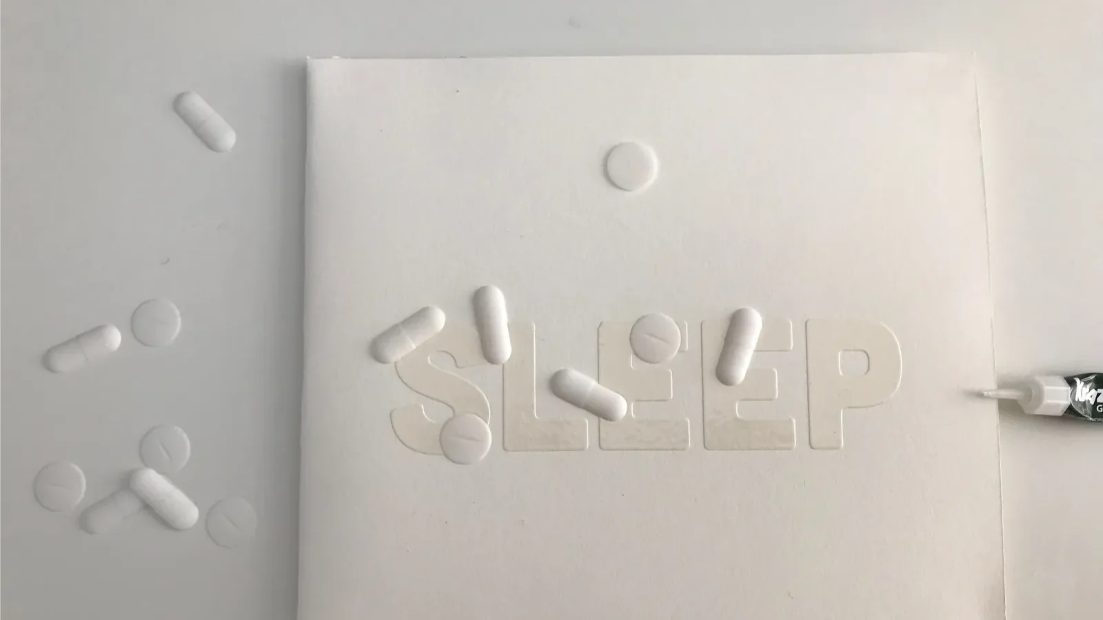 A SLEEP sleeve on a white table with 3D-printed pills glued over the glossy lettering, more pills beside it and a tube of glue.