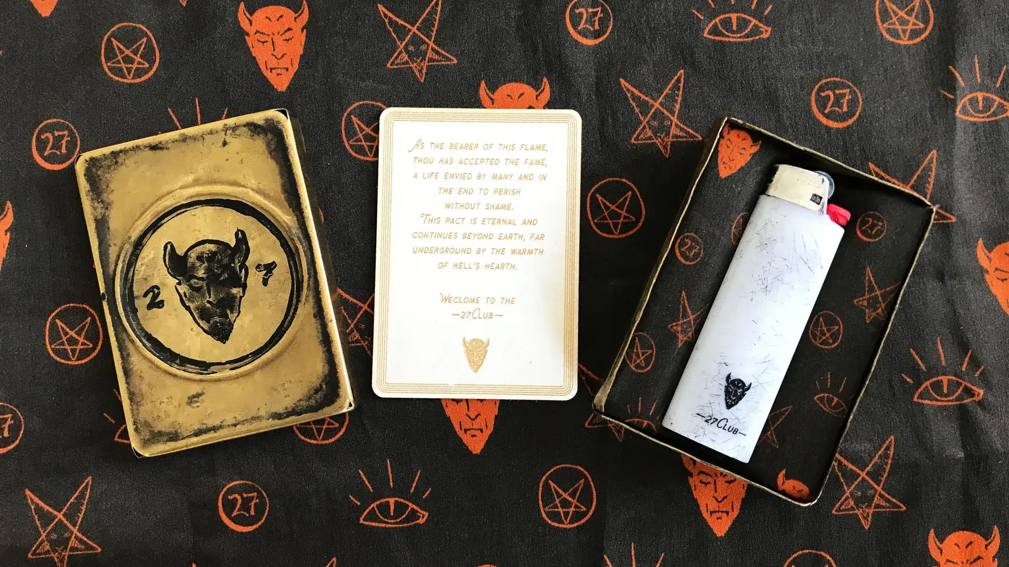 On black silk printed with orange devils, pentagrams and eyes: a worn brass case stamped with a devil and 27, a cream card of etched text, and the open case holding a scuffed white lighter.