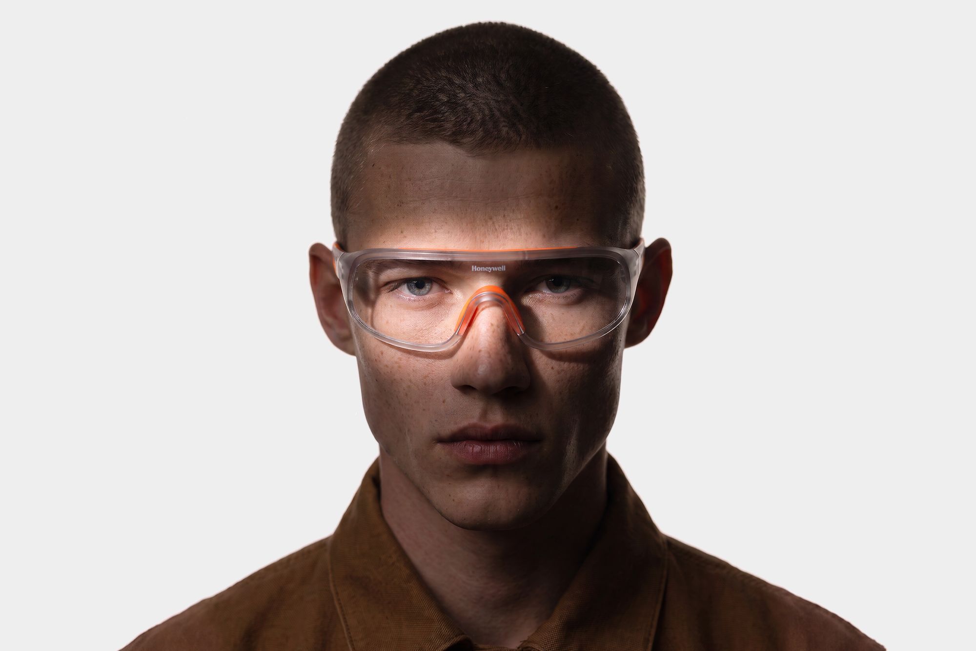 Portrait of a man in a brown work jacket wearing clear Honeywell safety glasses with an orange brow bar and nose bridge, lit from the side.