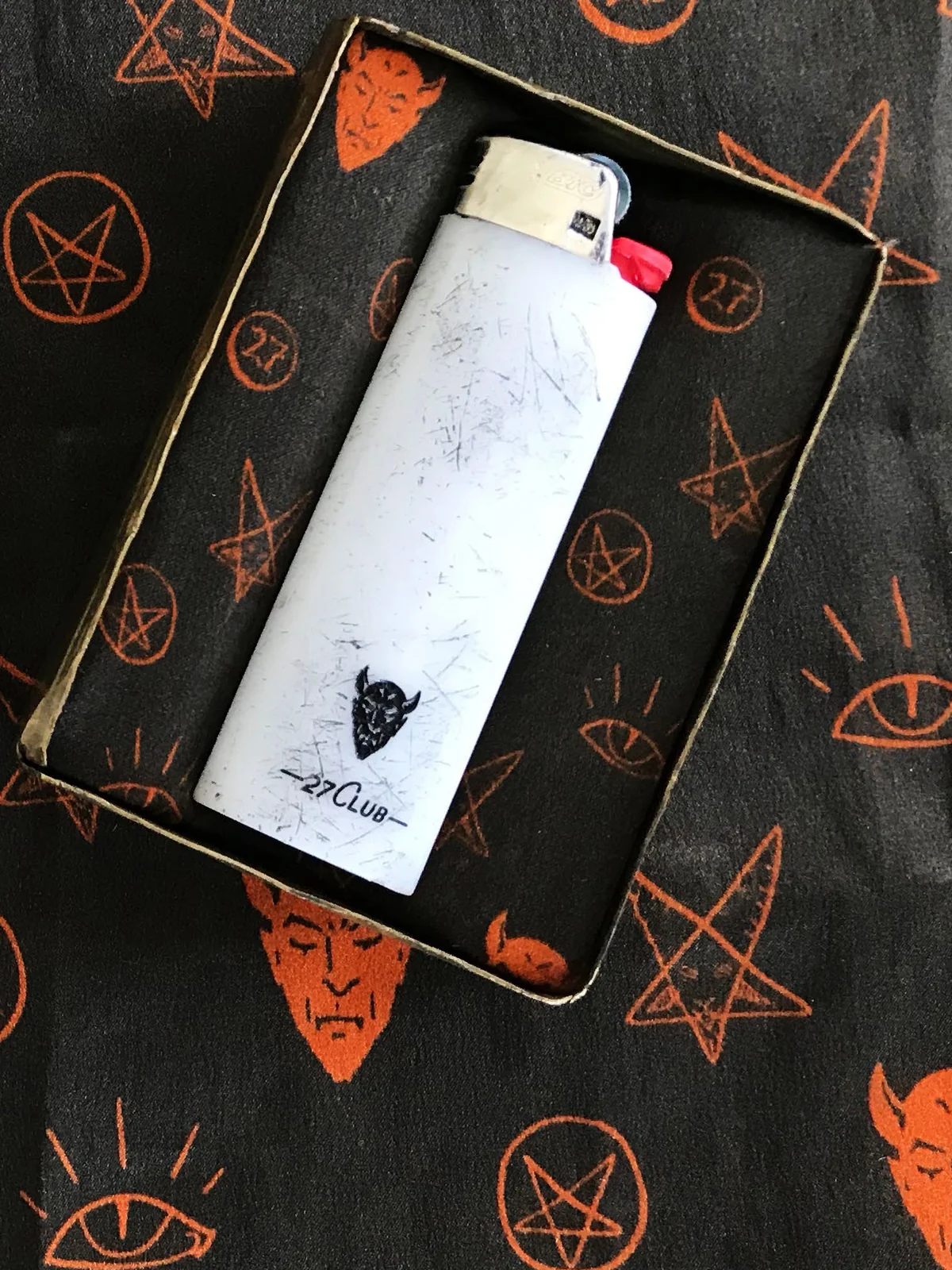 The open case, lined with black silk printed with orange devils, pentagrams and eyes, holding a scratched white lighter marked with a devil and 27 Club.