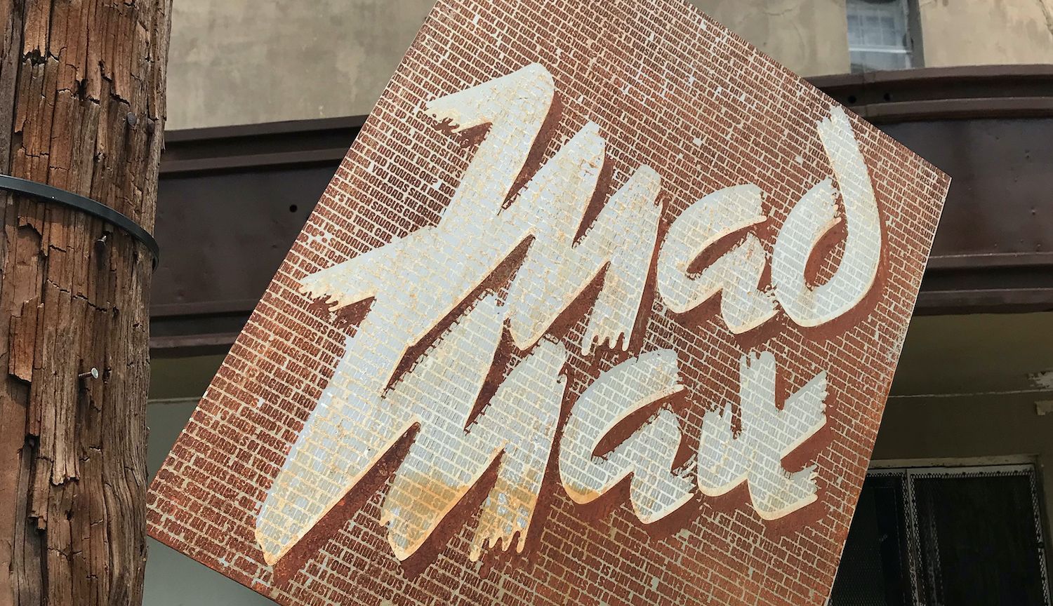 A rusted metal sign with Mad Max in brush lettering, mounted at an angle on a wooden utility pole.