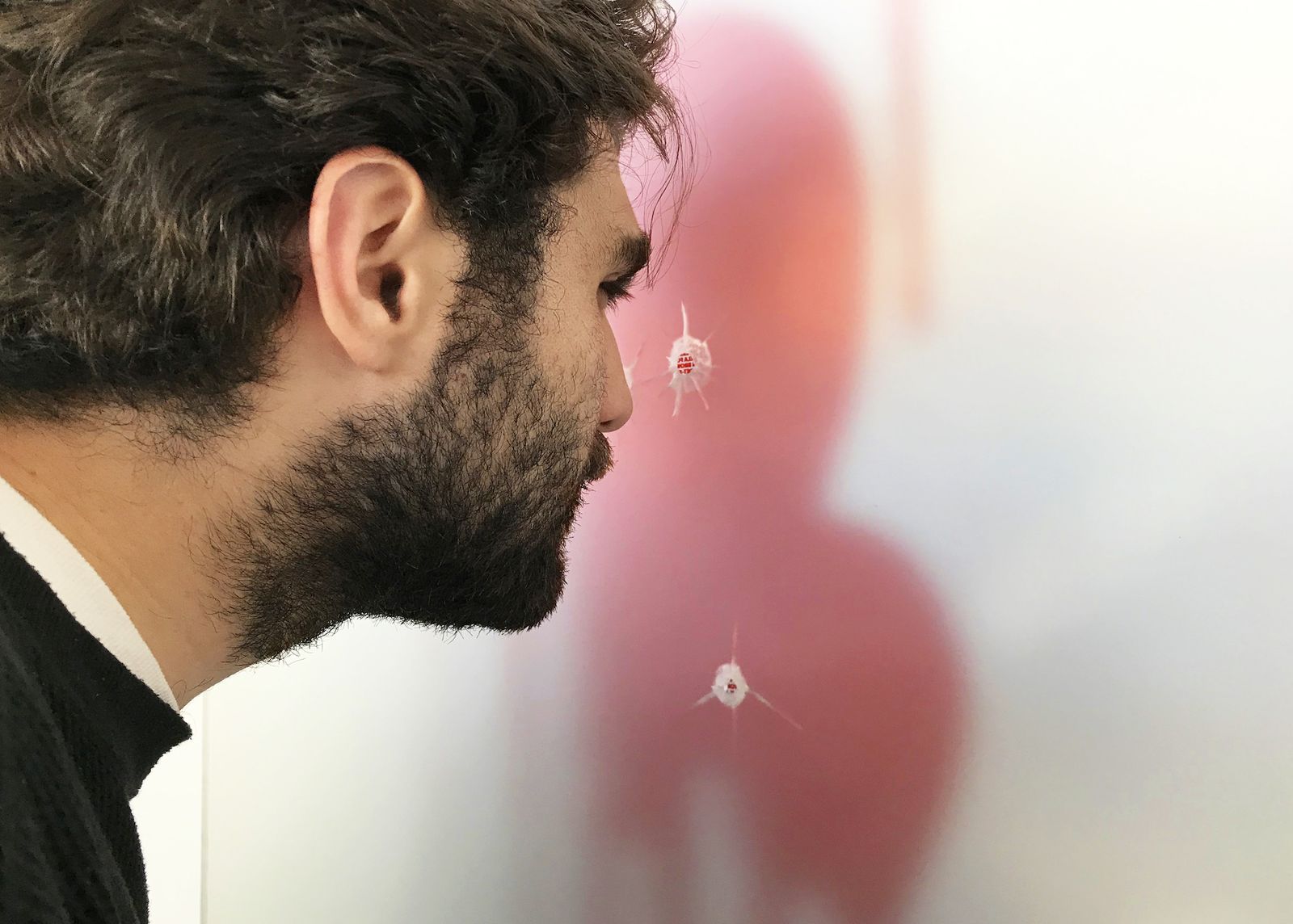 A man leans in close to a white panel to look at a blurred red human silhouette marked with bullet holes, each holding tiny printed text.
