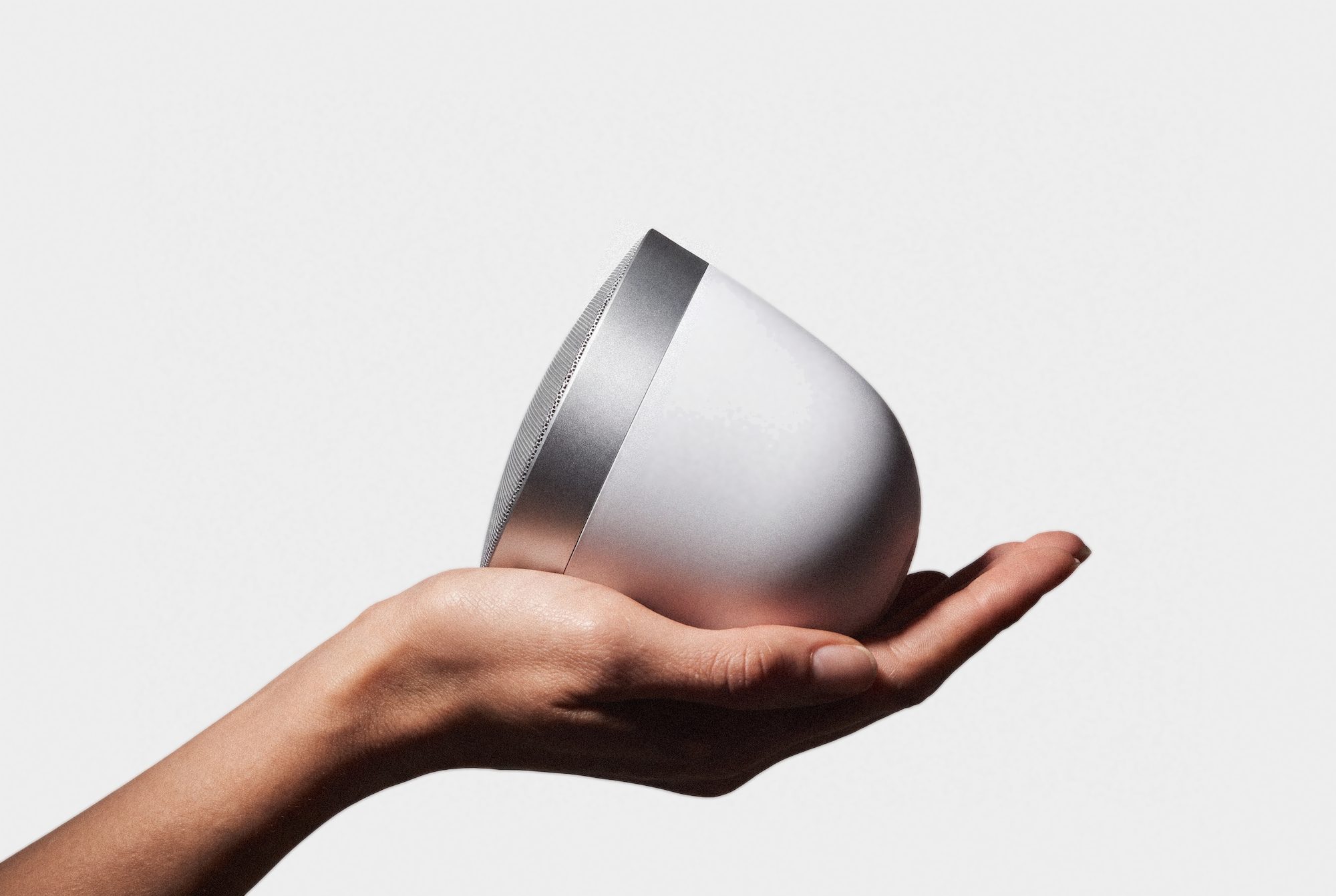 A hand holding the Kohler Moxie 2 speaker: a white rounded body with a metal band and a perforated speaker grille on its face.