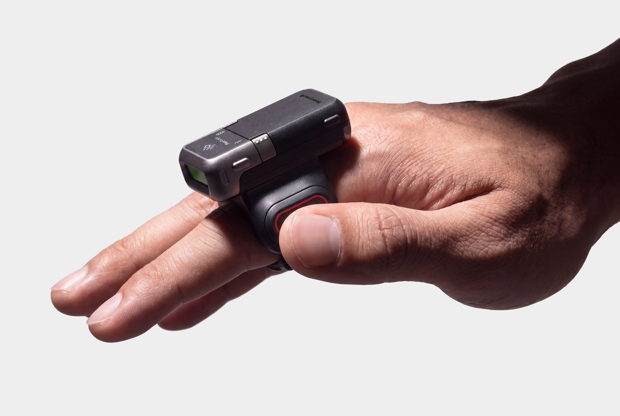 A hand wearing a compact dark gray Honeywell ring scanner on the index finger, the thumb pressing its red-ringed trigger and the green scan window facing forward.