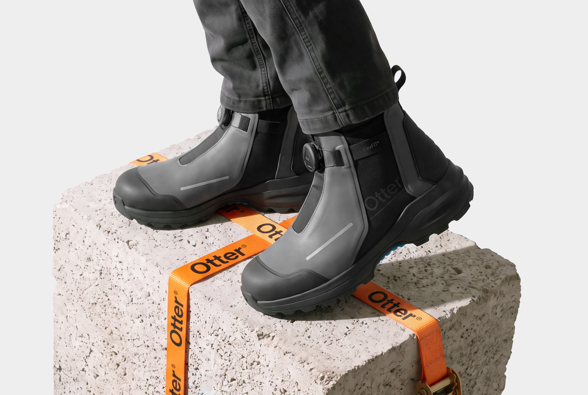 A person in dark work jeans standing on a rough concrete block in a pair of gray and black Otter safety boots with dial closures, an orange Otter strap ratcheted around the block.