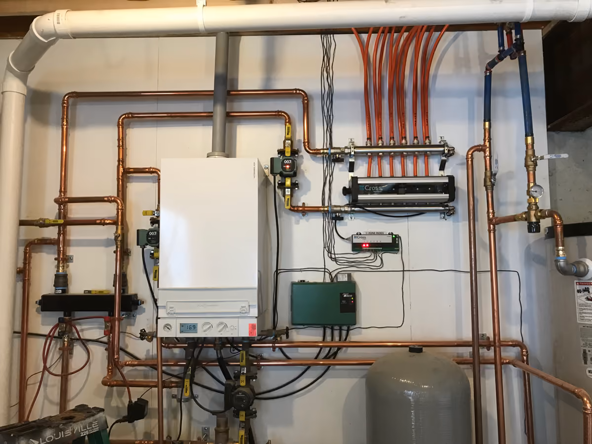 Indoor plumbing system with copper pipes, a white boiler, red tubing, control panels, and a gray pressure tank.