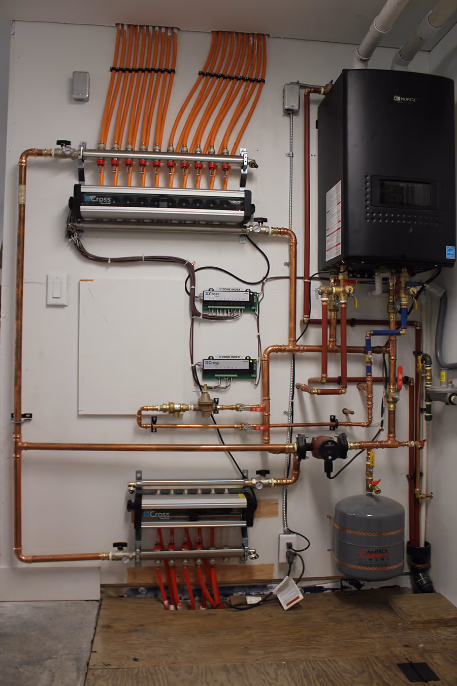 Wall-mounted plumbing and heating system with copper pipes, valves, orange and red tubing, and a black Noritz unit.