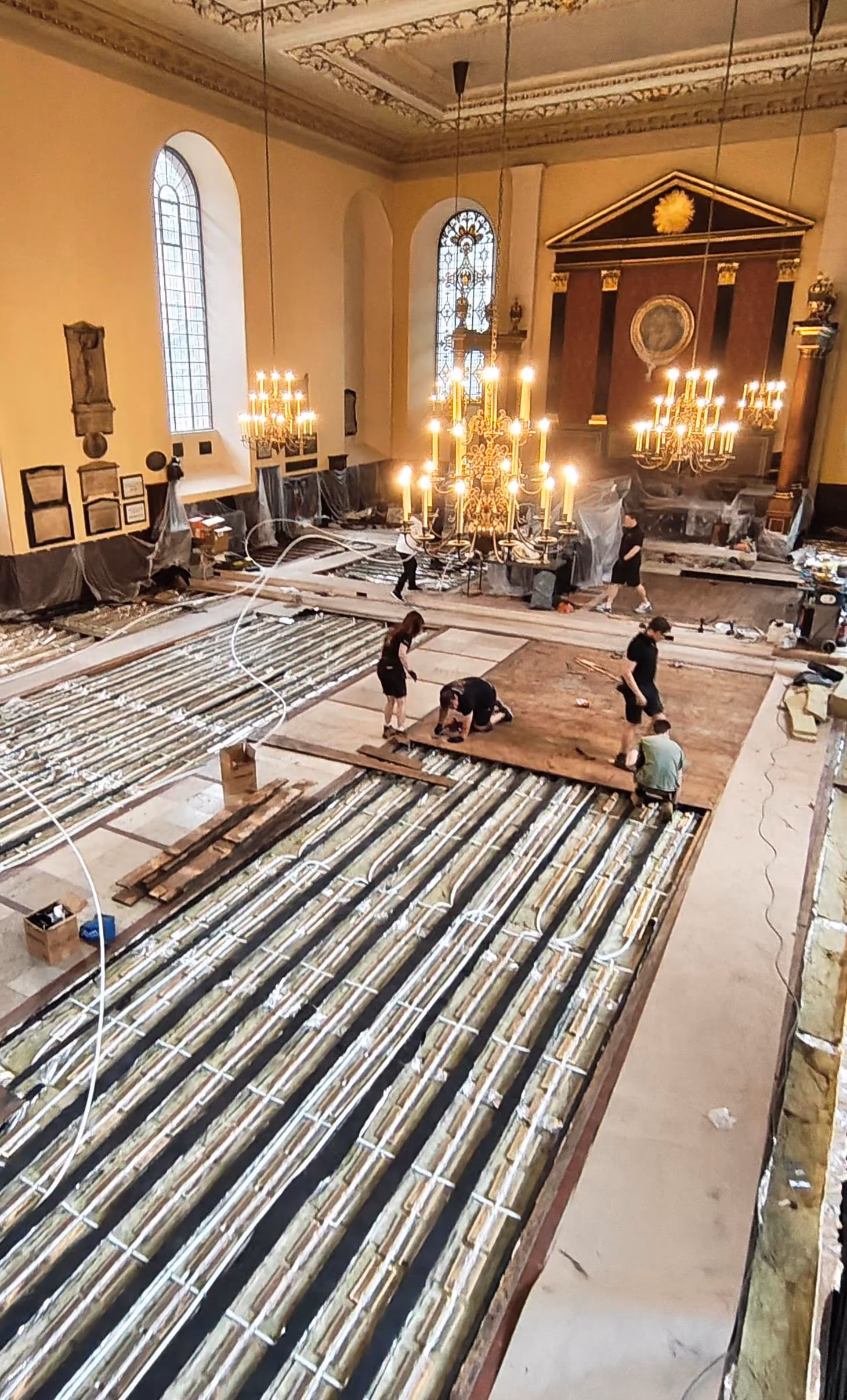 Workers installing or repairing flooring with visible insulation and wiring inside a large, chandelier-lit hall.