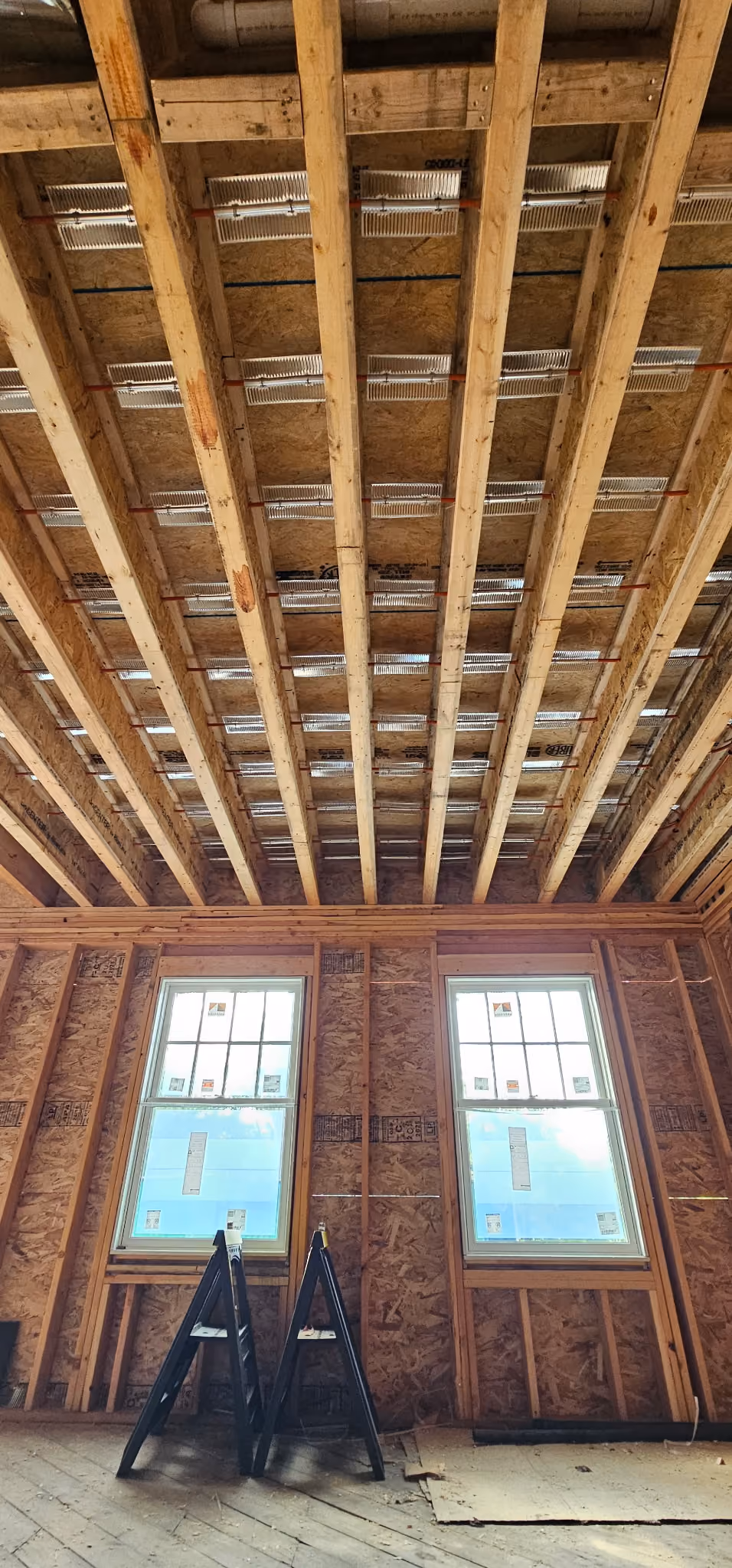 Interior of a wooden-framed room under construction with exposed ceiling joists, oriented strand board walls, two large windows, and two black sawhorses on a wooden floor.