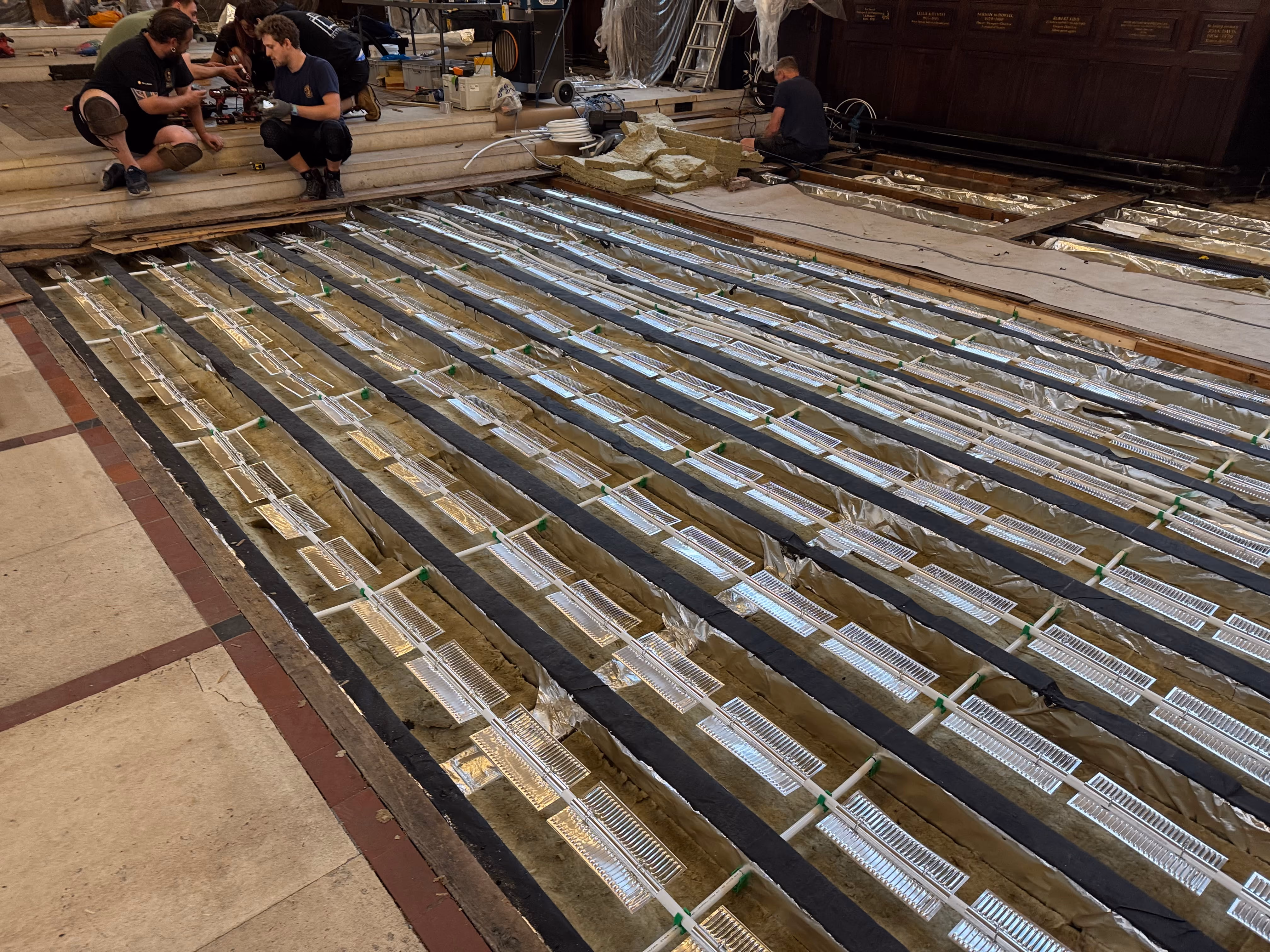 Construction workers installing underfloor heating system with metal heat spreaders and pipes exposed on a large floor inside a building under renovation.