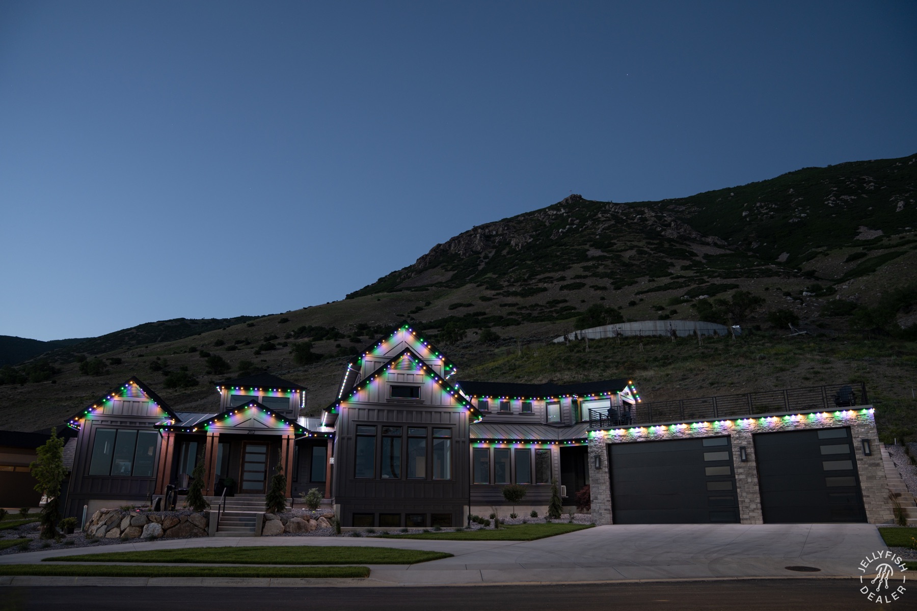 Purple, green and orange roofline lights on a modern farmhouse beneath Utah foothills.