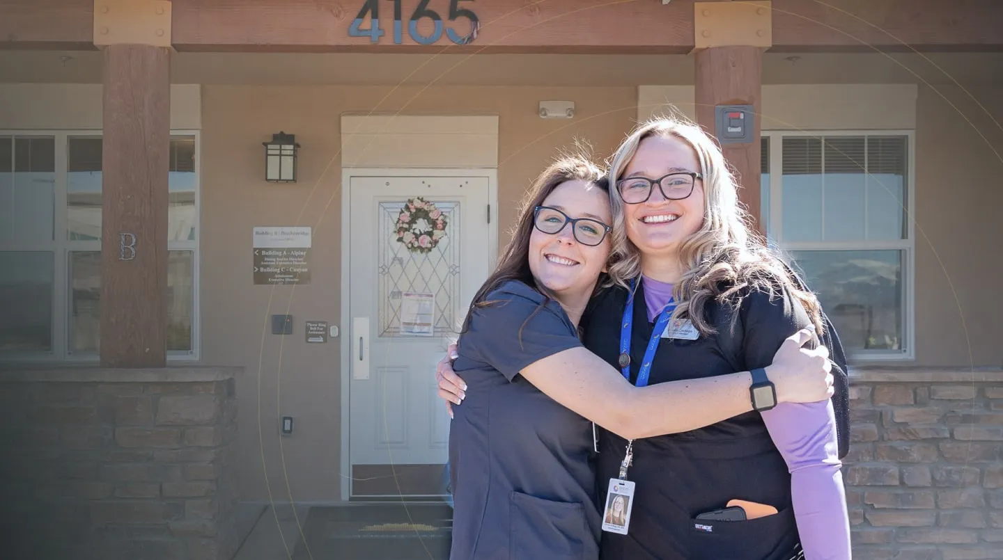Two smiling women in medical scrubs hugging in front of building entrance with the number 4165 above.