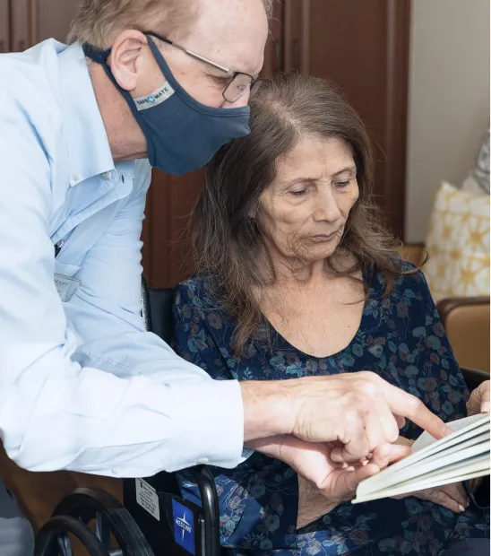 Man wearing mask points to book while seated woman looks on attentively.
