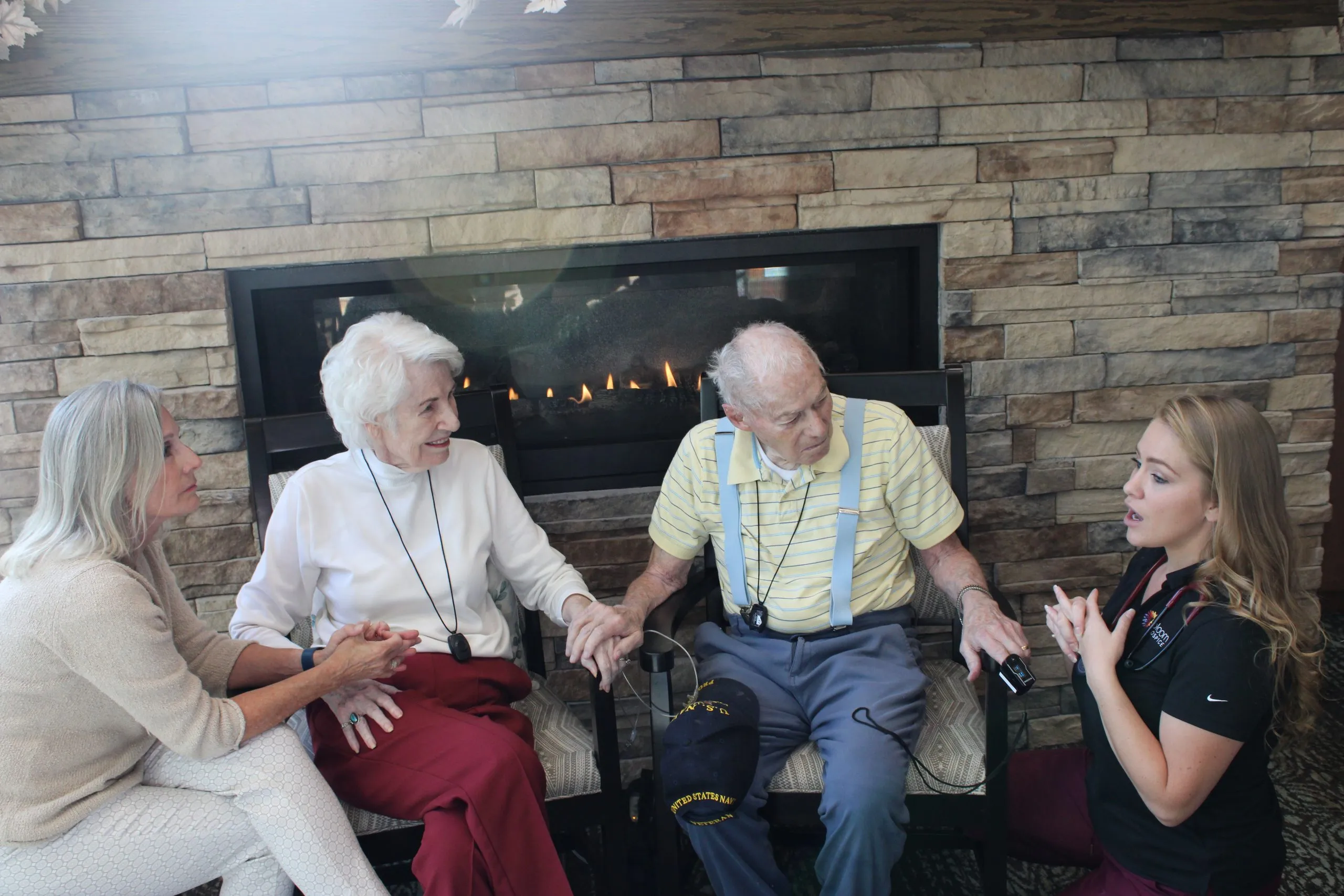 Elderly couple holding hands talk with a seated nurse by a stone fireplace in a cozy room.