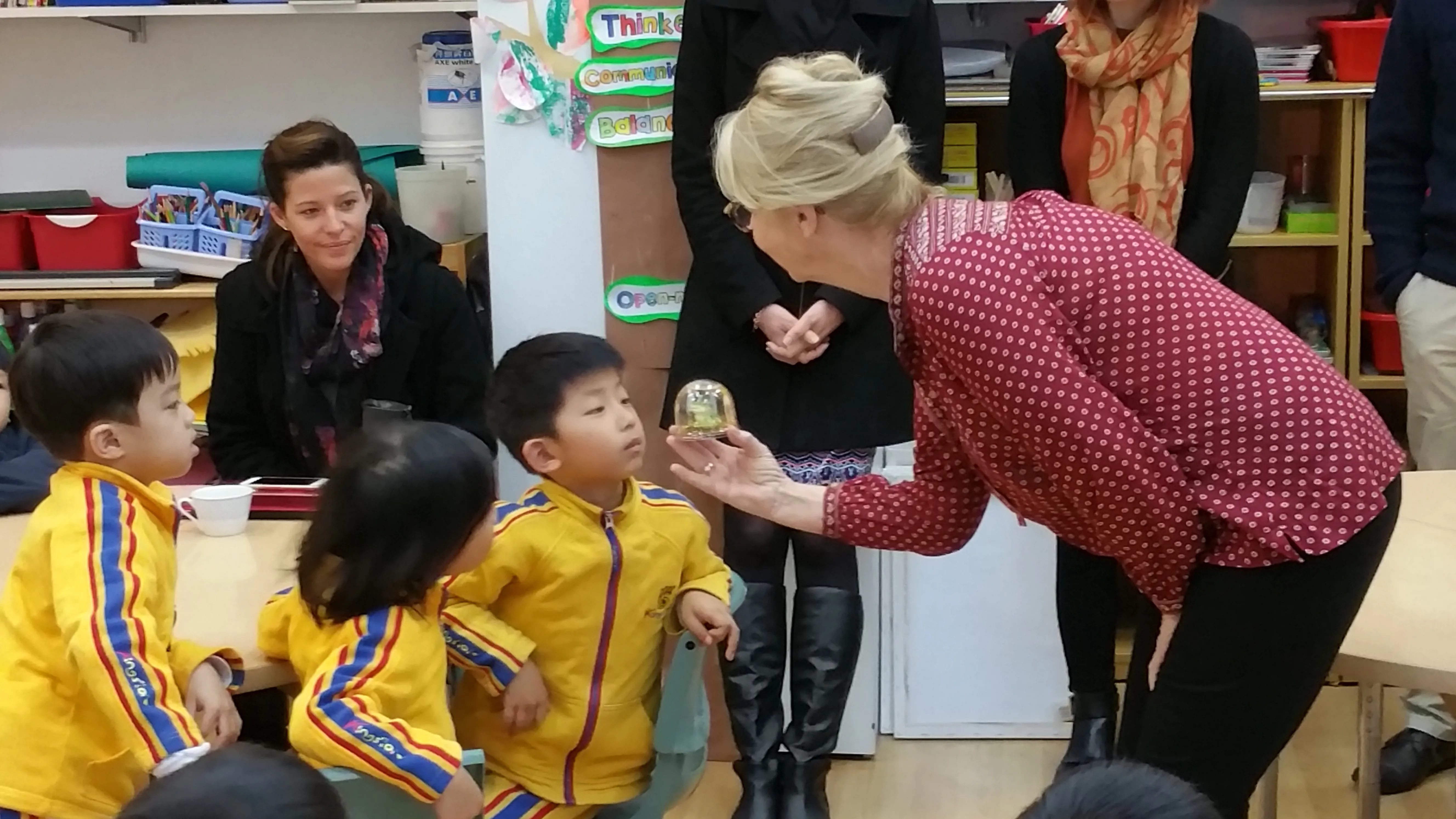 A woman in a red patterned shirt bends forward showing a small object in a transparent dome to young children in yellow jackets sitting at tables in a classroom.