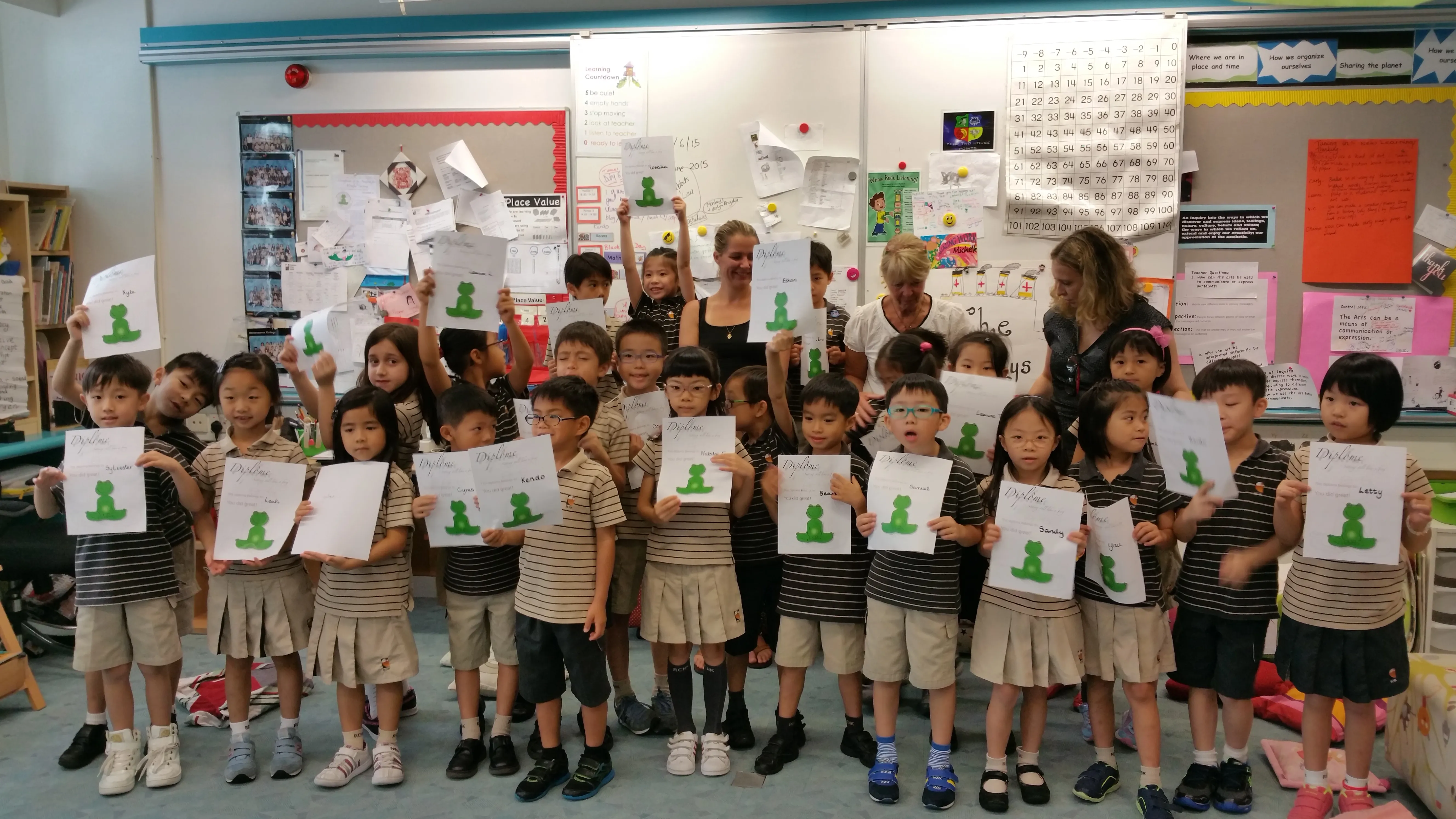 A group of young school children in uniform standing in a classroom holding up diplomas with a green frog image, smiling and celebrating their achievement, with three adults standing behind them.