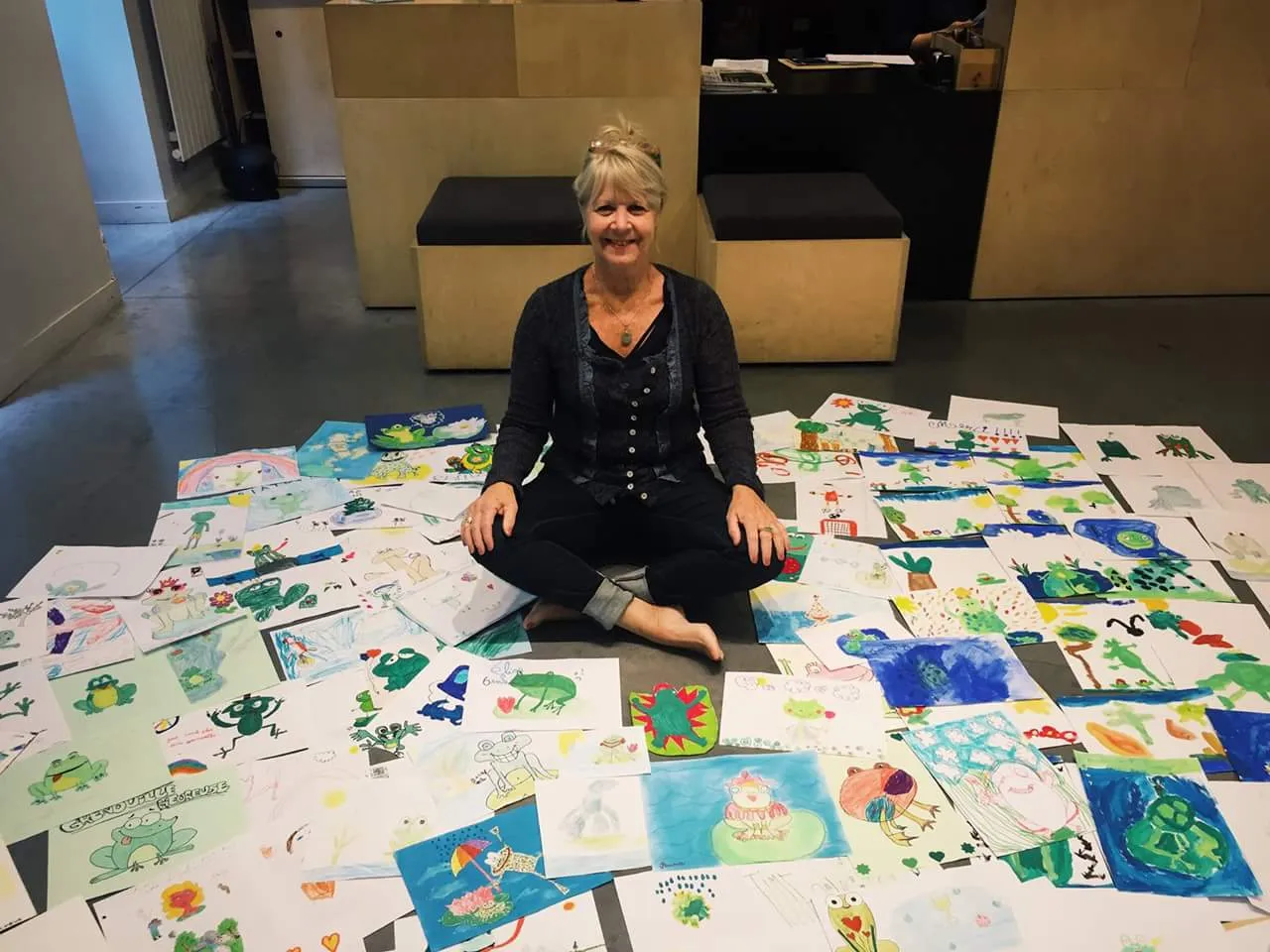 Woman sitting cross-legged on the floor surrounded by numerous colorful drawings of frogs and other imaginative creatures.