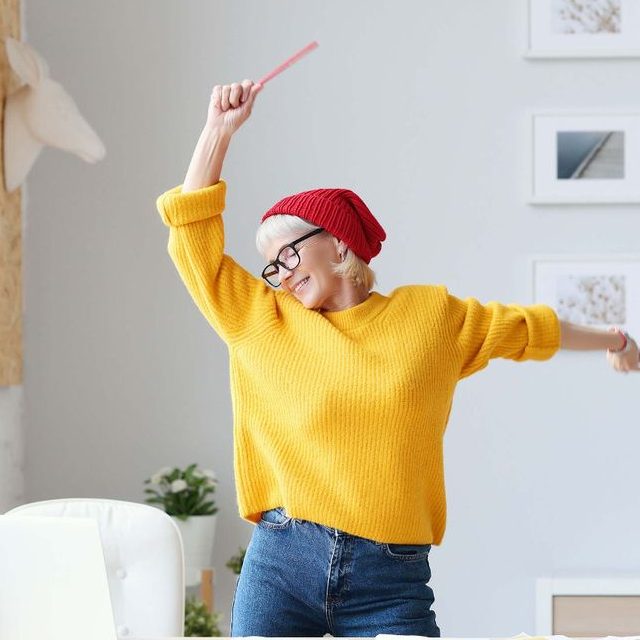 Smiling elderly woman with glasses, wearing a red beanie and yellow sweater, joyfully dancing indoors with one arm raised holding a pink object.
