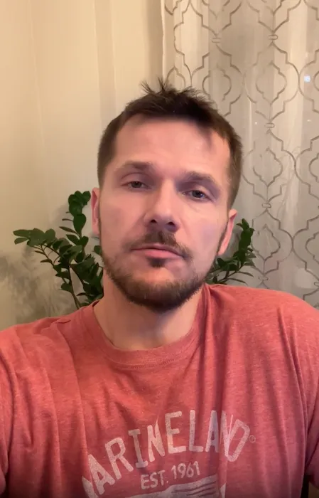 A man with short brown hair and a beard wearing a red T-shirt with the word 'MARINELAND' printed on it, sitting indoors with a plant and patterned curtains in the background.