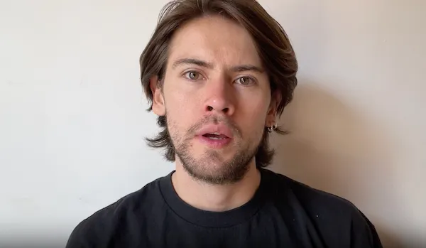 Young man with medium-length brown hair and light facial hair, wearing a black shirt, looking directly at the camera against a plain light background.