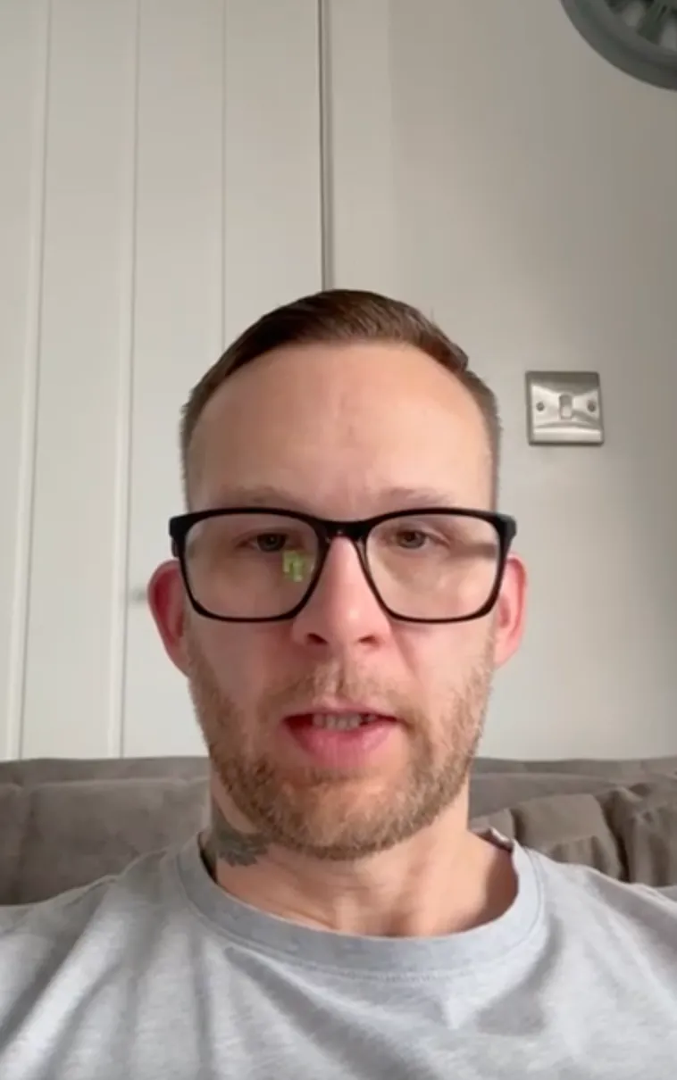 Man with short brown hair, glasses, and beard wearing a grey t-shirt sitting on a grey sofa with a white paneled wall and a light switch behind him.