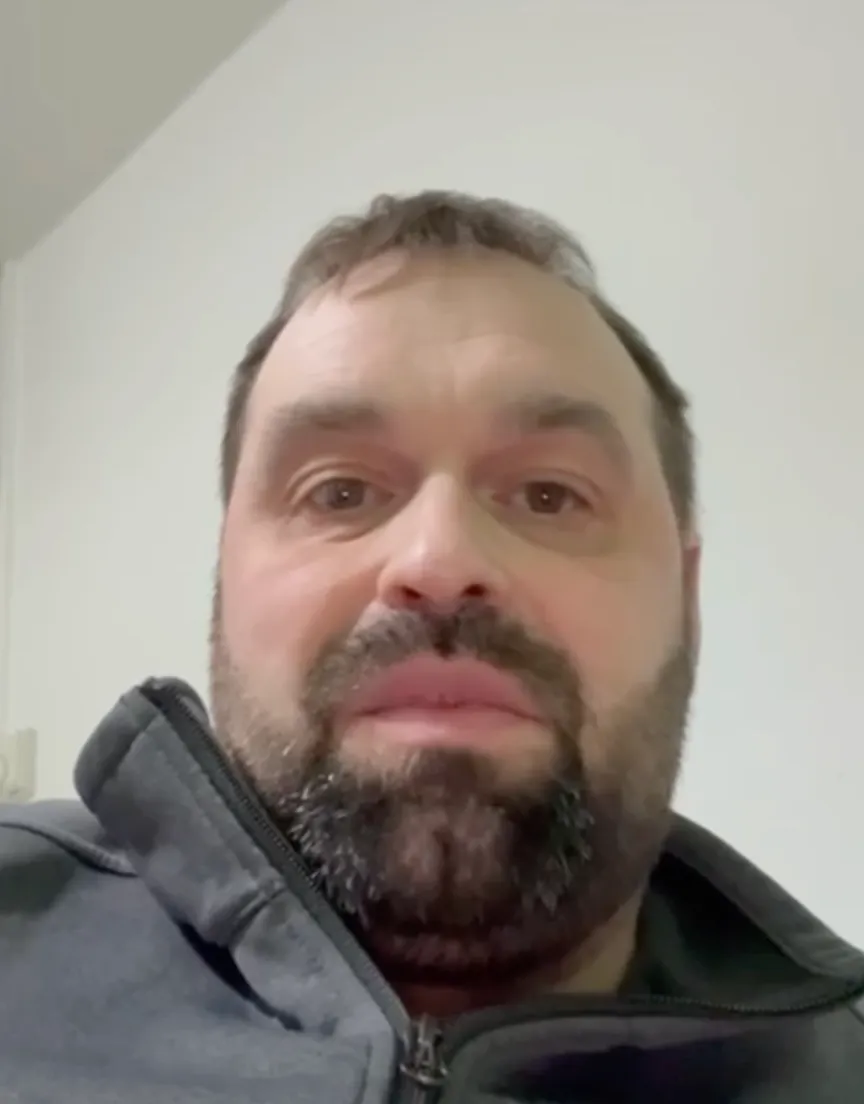 Close-up of a man with short dark hair and a thick beard wearing a zipped-up dark grey jacket against a plain white wall background.