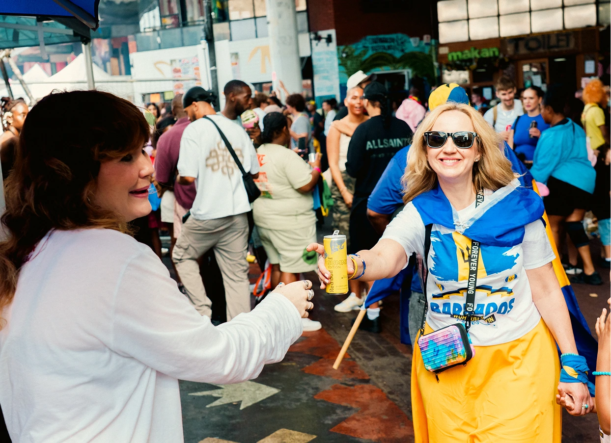 A smiling woman wearing sunglasses, a yellow skirt, and a Barbados flag draped over her shoulders hands a can of Red Bull Summer Edition to another woman in a white shirt, with a crowd of people and event decorations in the background.