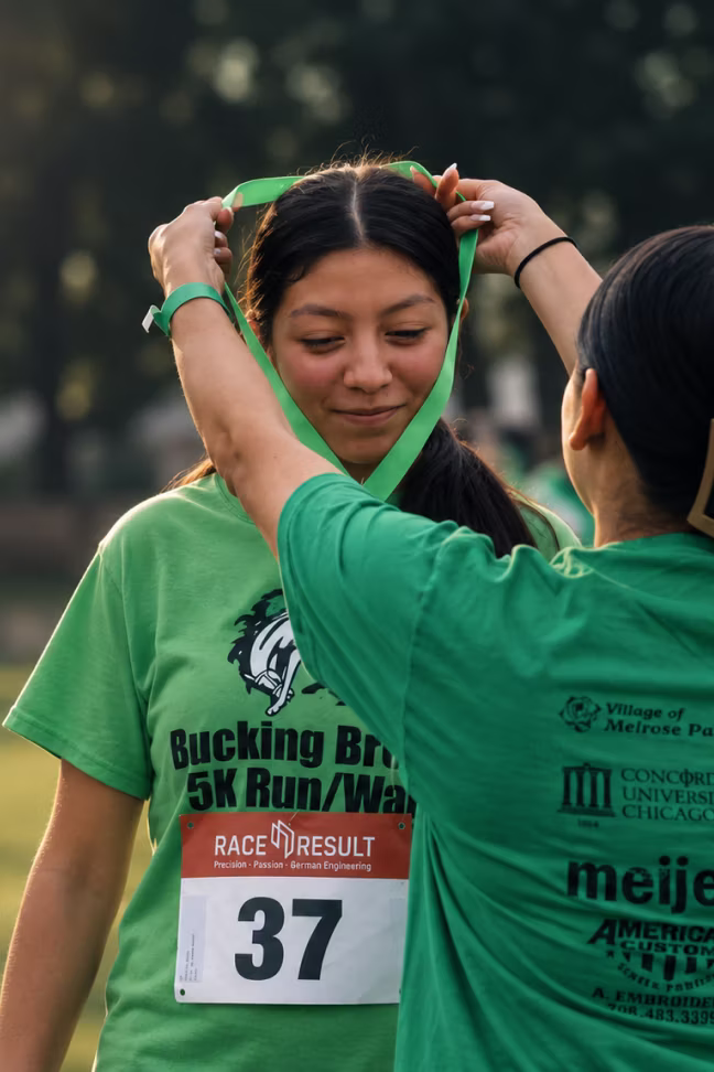 A young woman wearing a green Bucking Bronco 5K Run/Walk t-shirt with race bib number 37 is receiving a green medal from another person dressed in green outdoors.