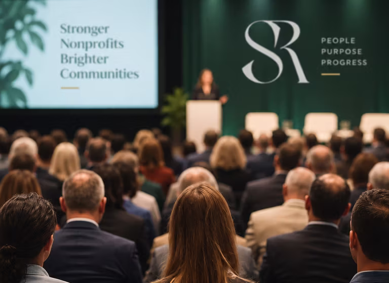 Audience seated facing a stage where a speaker is presenting in a conference room with banners, one reading 'Stronger Nonprofits Brighter Communities' and another showing the initials 'SR' with the words 'People Purpose Progress'.