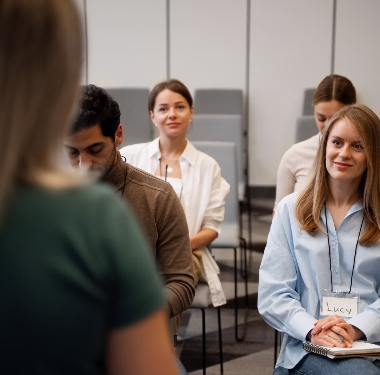 A group of people seated in chairs listening attentively to a speaker whose back is to the camera; a woman in the front right wears a name tag labeled Lucy and holds a notebook.