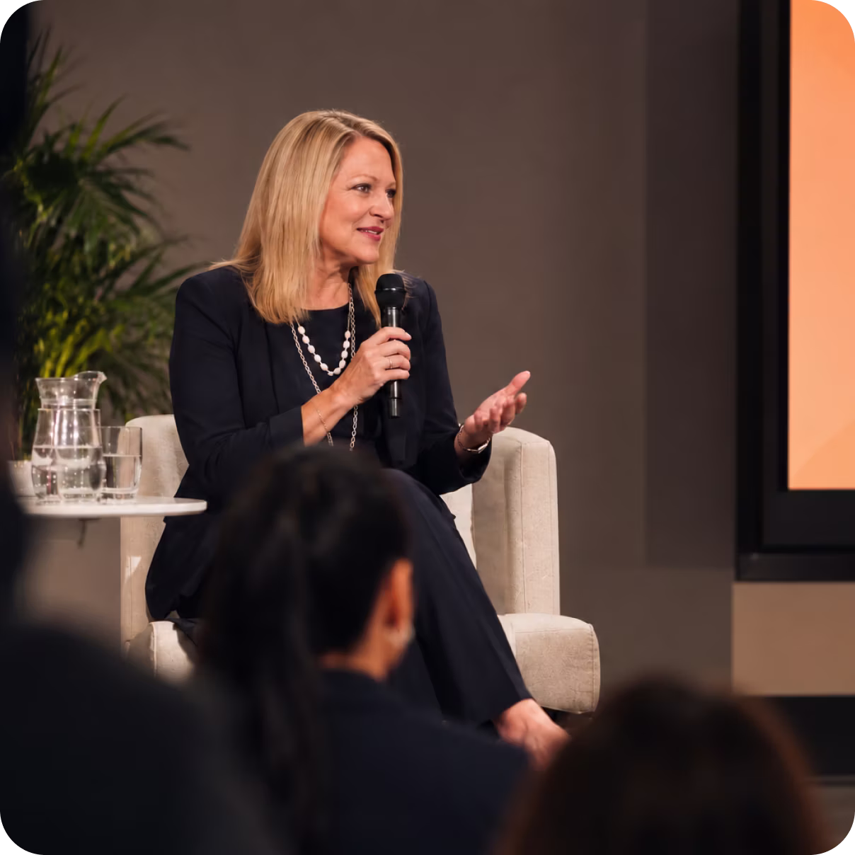 A blonde woman in a black suit sitting on a beige chair speaking into a microphone with a glass pitcher and glass of water on a table next to her during a discussion or event.