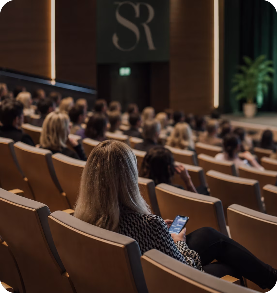 Audience seated in rows of beige chairs at a conference or event, with a woman in the foreground looking at her phone, a stage with a potted plant visible in the background.