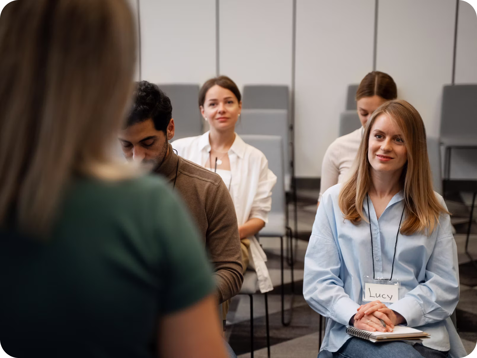A group of four people seated in a classroom or seminar setting, with one woman in the foreground facing them; a smiling woman named Lucy wearing a name tag sits closest to the camera holding a notebook.