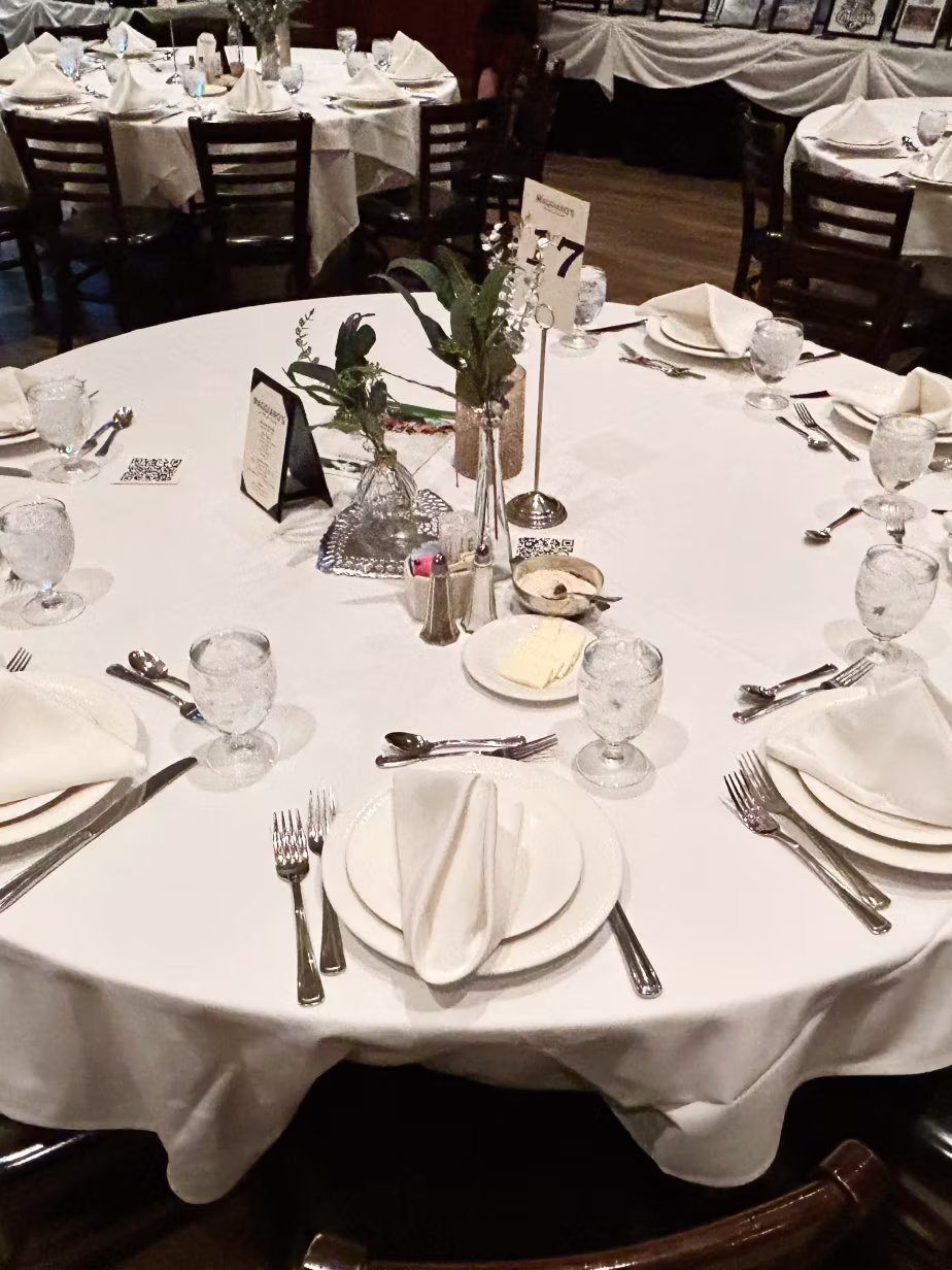 Round restaurant table set for six with white tablecloth, folded napkins on plates, glass goblets, silverware, butter dish, salt and pepper shakers, small floral centerpieces, and a table number 7 sign.