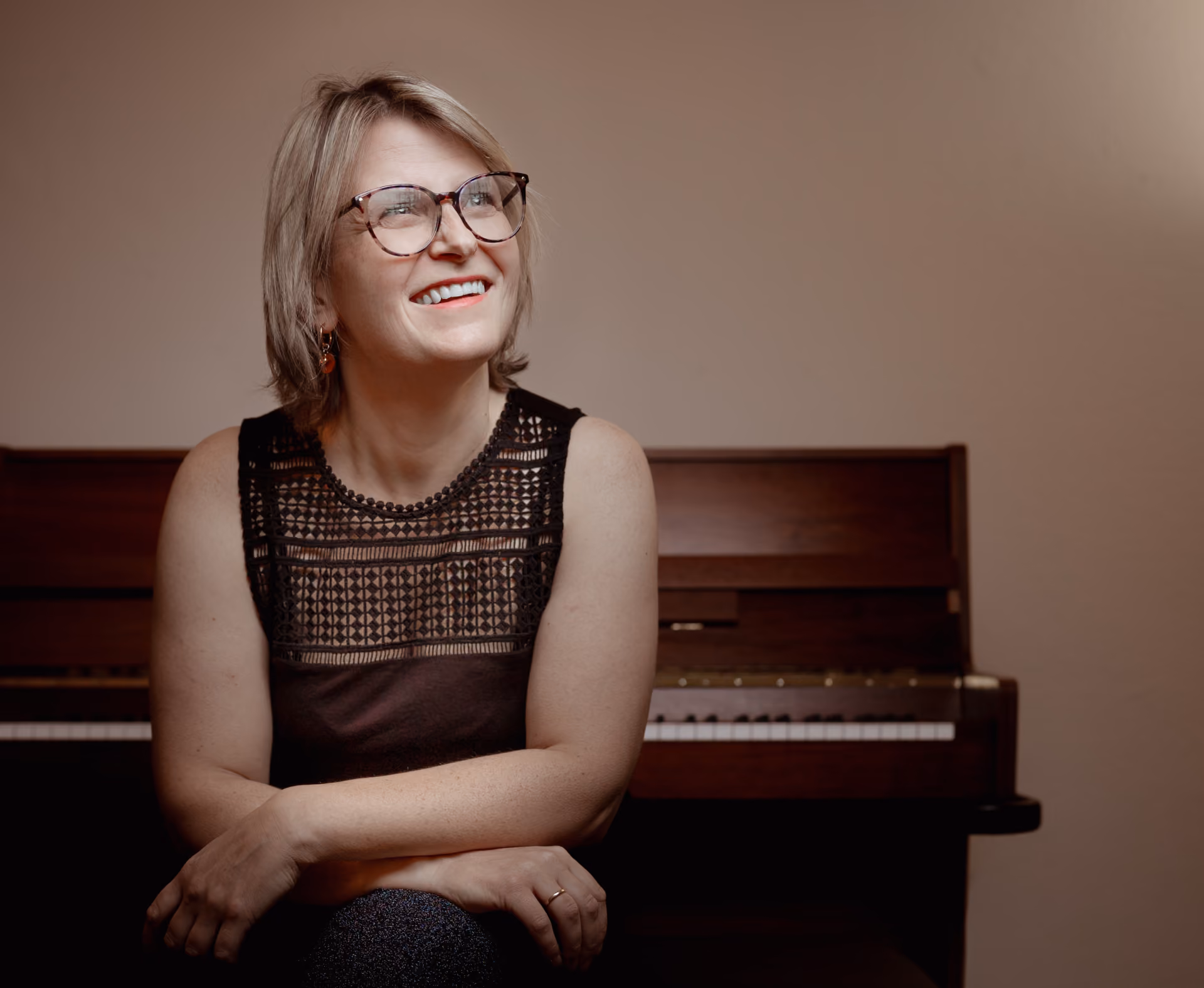 Frau mit Brille und schwarzem Spitzenoberteil sitzt vor einem Klavier und lächelt nach rechts oben.