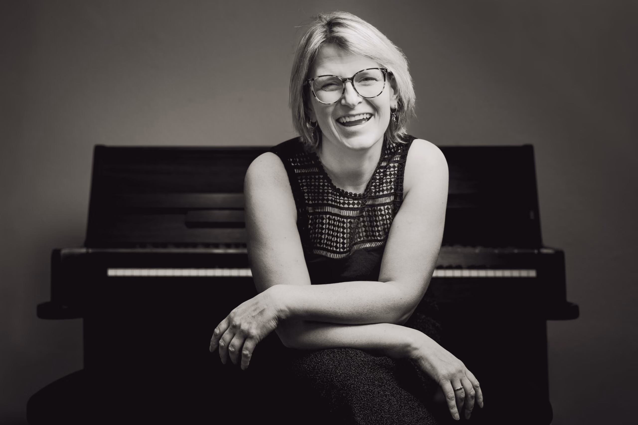 Schwarz-weiß-Foto einer lächelnden Frau mit kurzen blonden Haaren und Brille, die vor einem Klavier sitzt, die Arme verschränkt.