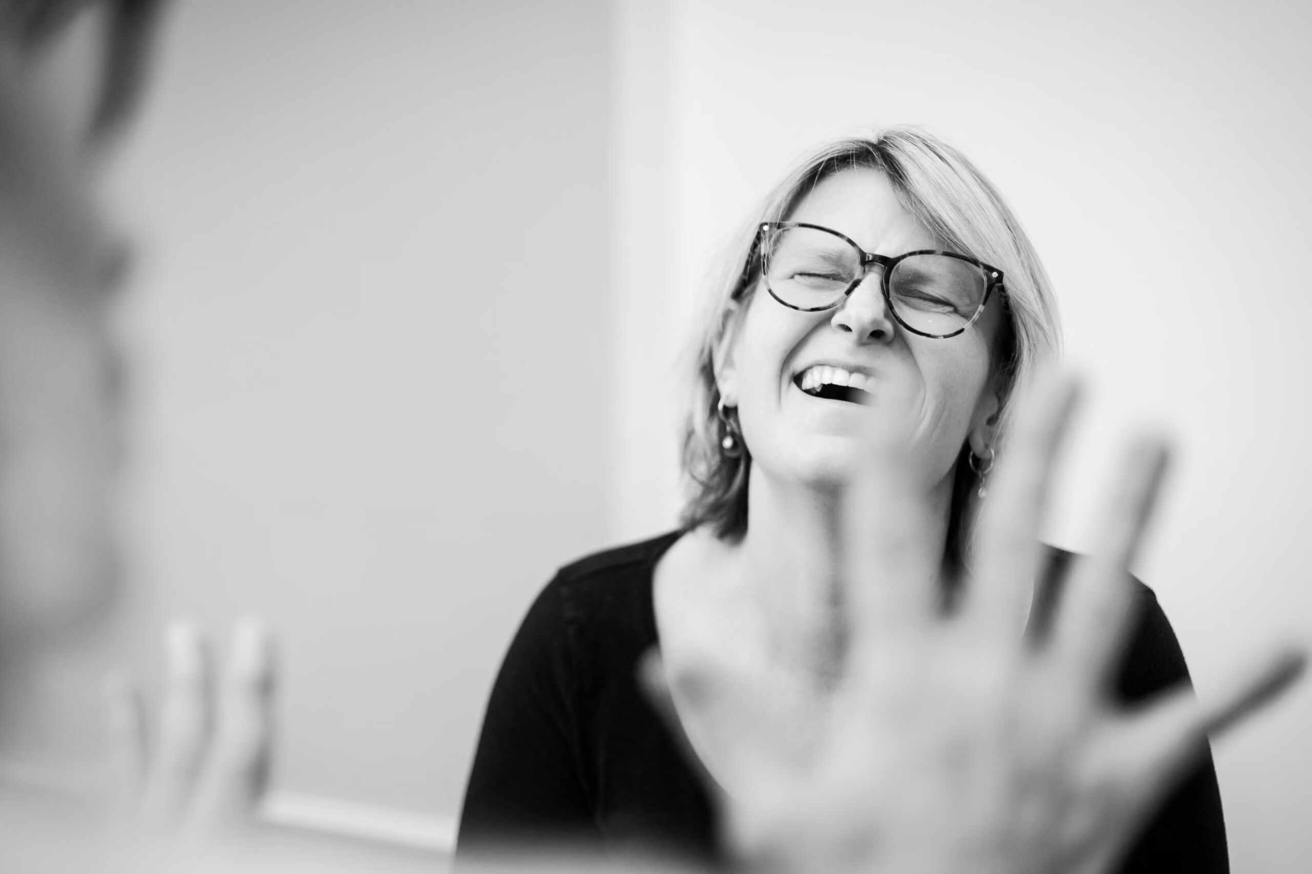 Schwarz-weiß-Foto einer Frau mit Brille, die mit geschlossenen Augen und offenem Mund lacht, vor einer verschwommenen Hand im Vordergrund.