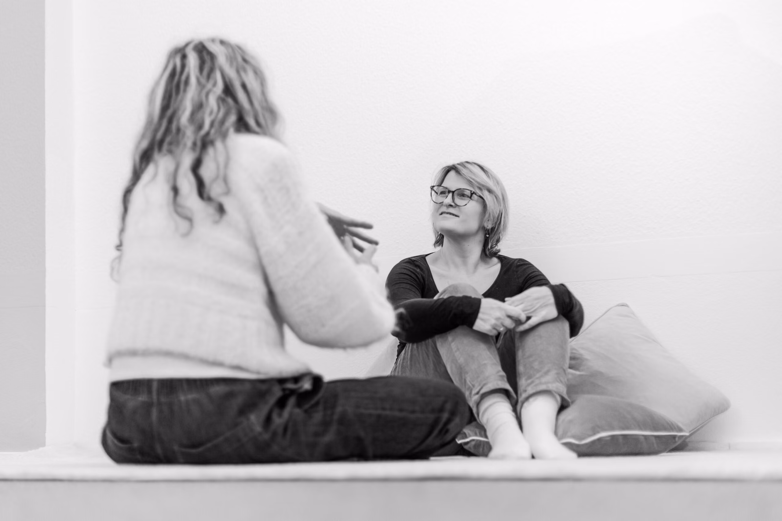 Zwei Frauen sitzen sich in einem hellen Raum gegenüber, eine spricht, während die andere aufmerksam zuhört und auf einem Kissen lehnt.