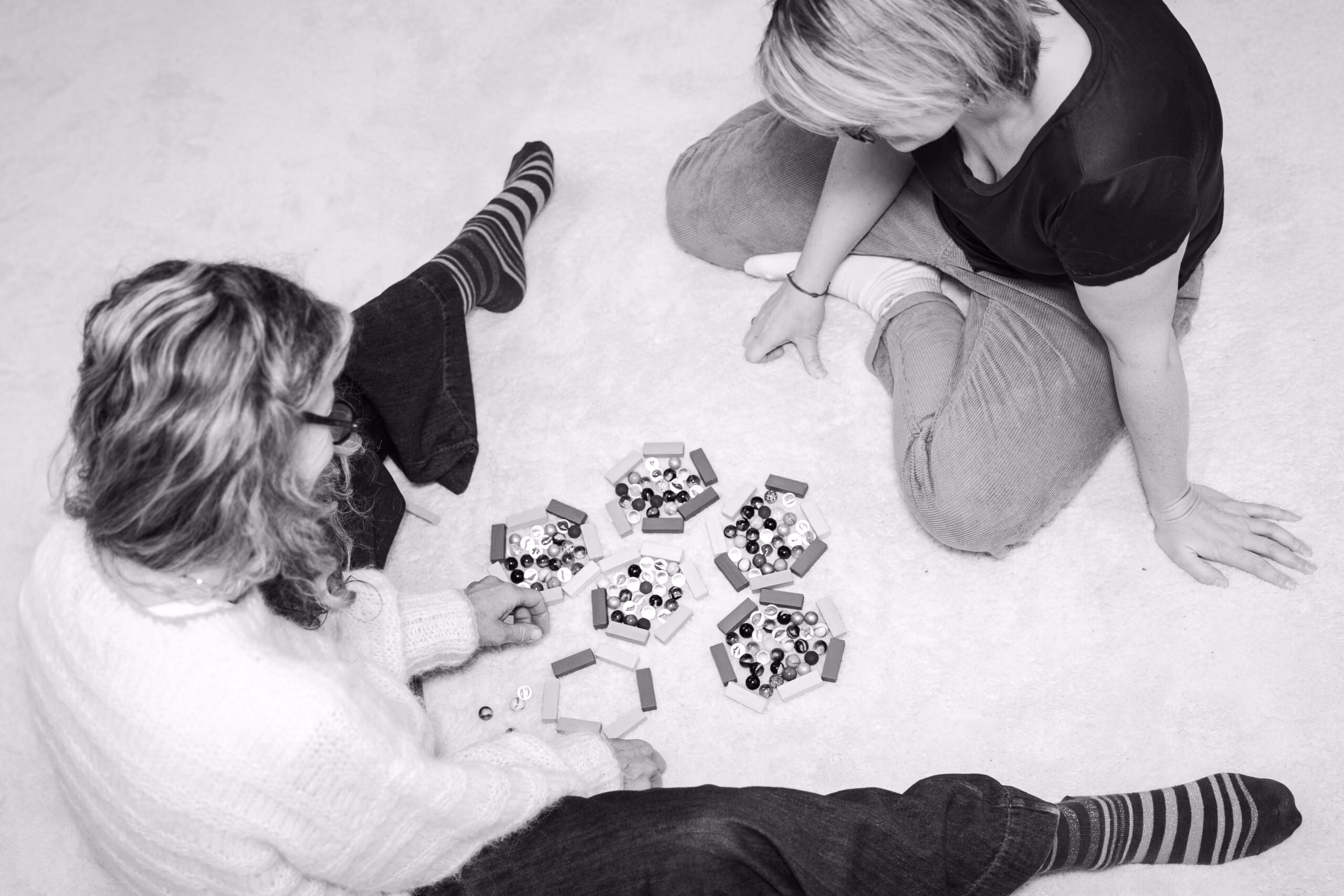 Zwei Frauen sitzen auf dem Boden und spielen ein Brettspiel mit bunten kleinen Kugeln und rechteckigen Spielsteinen.