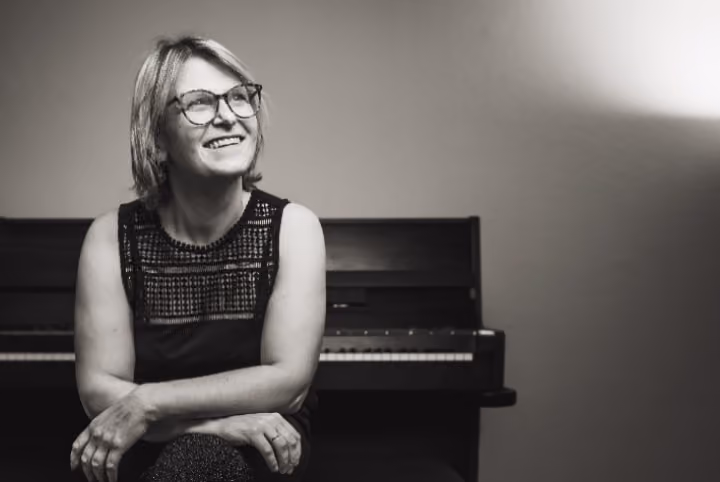 Schwarz-weiß-Porträt einer lächelnden Frau mit Brille, die vor einem Klavier sitzt und nach links schaut.